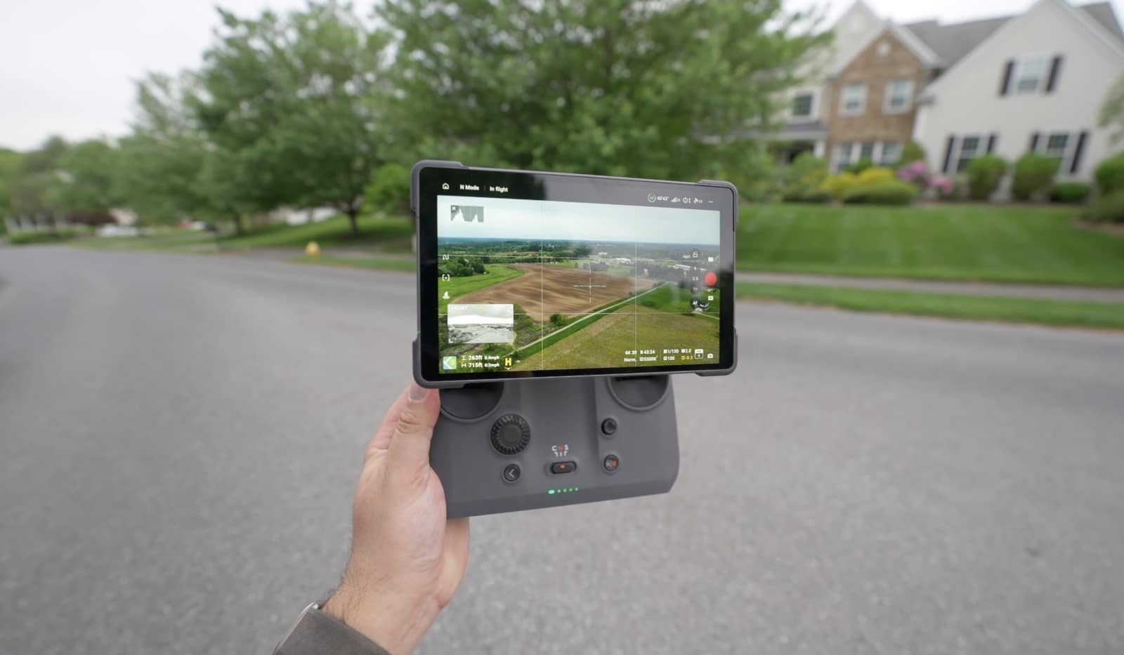 A hand raises a screen-equipped DJI controller showing live view on a suburban street.
