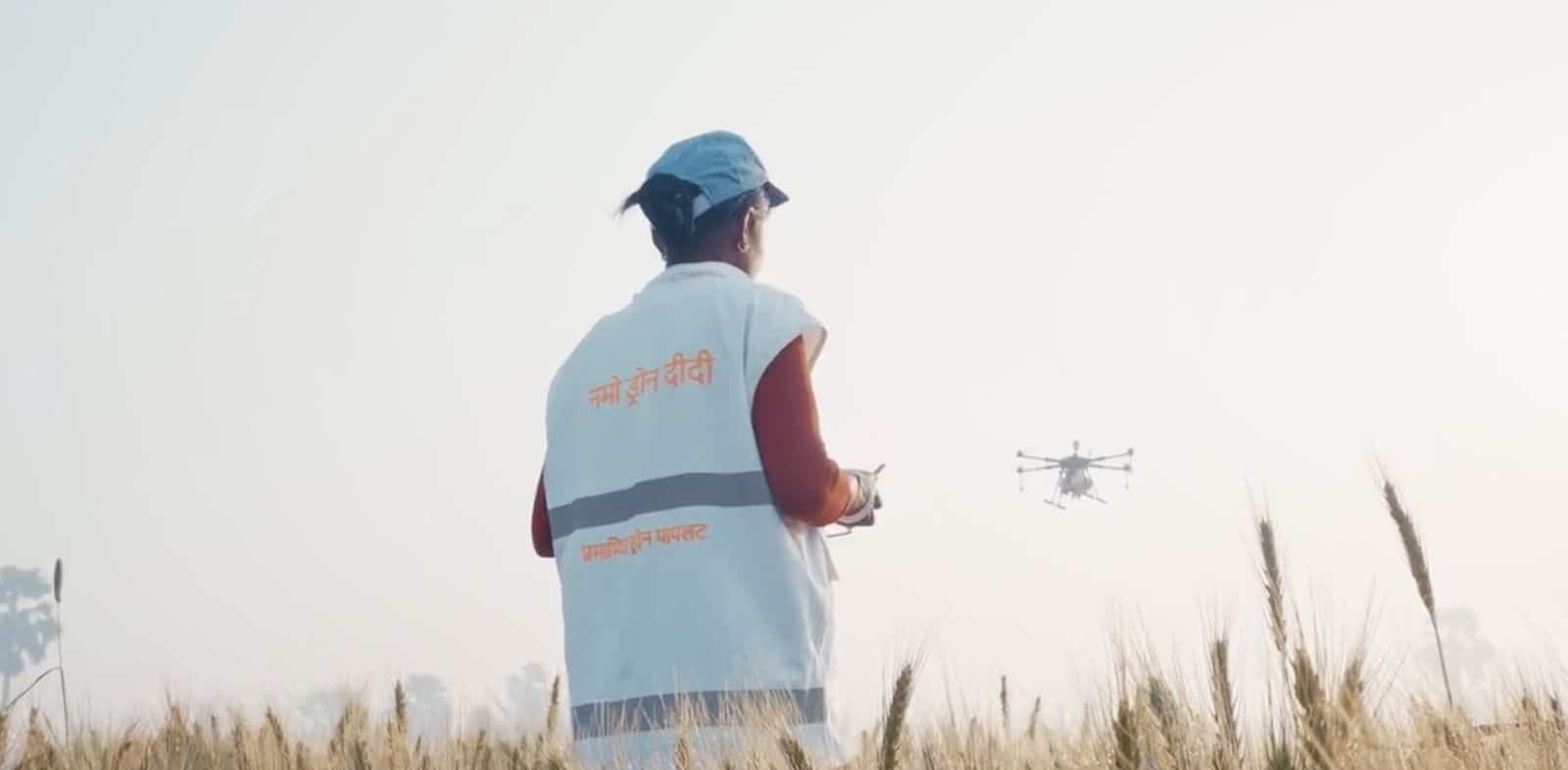 A woman in a marked white vest pilots a spray drone over a field.