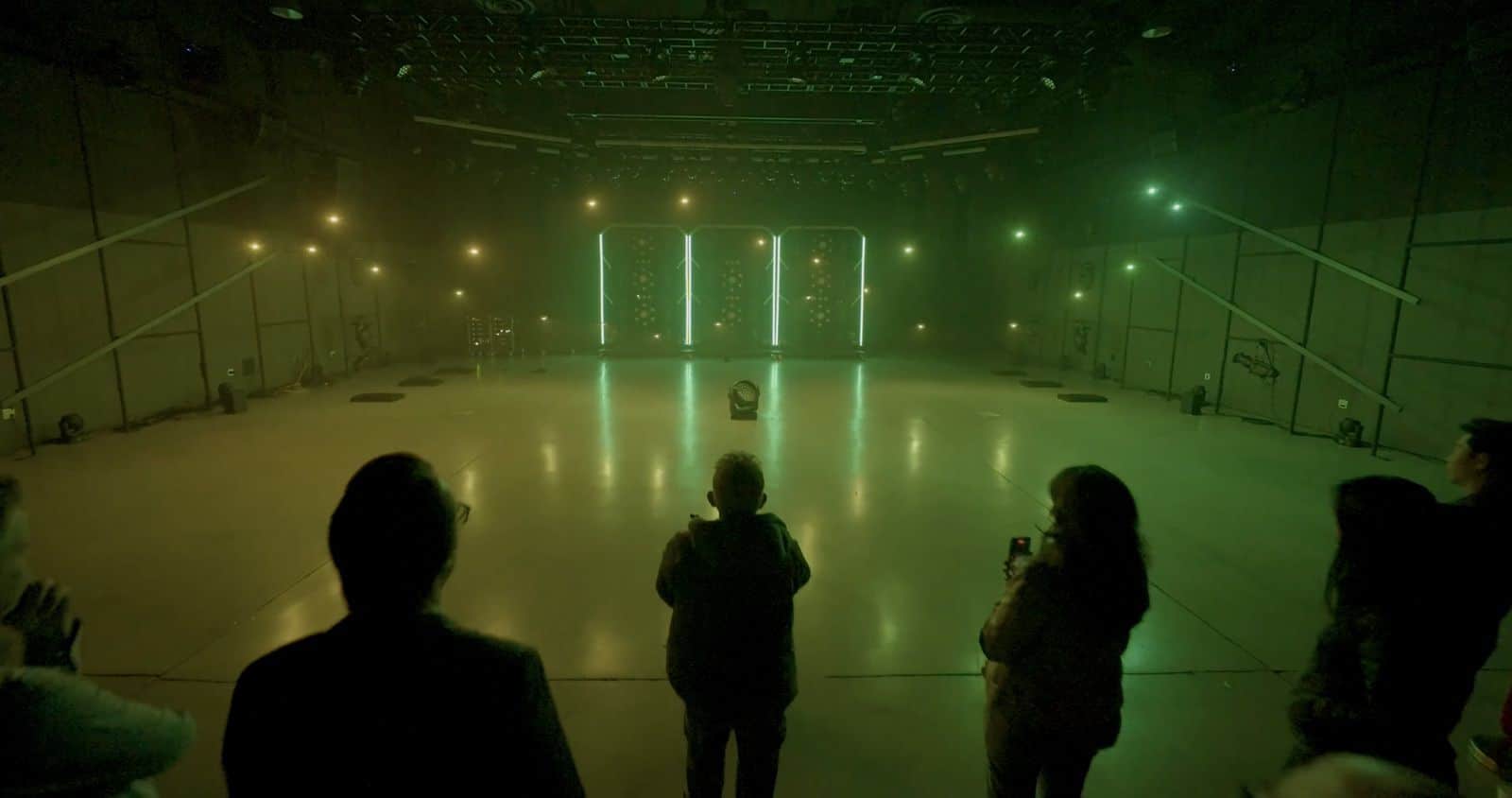 Green beams and hovering drones fill a hazy studio as spectators watch in silhouette.
