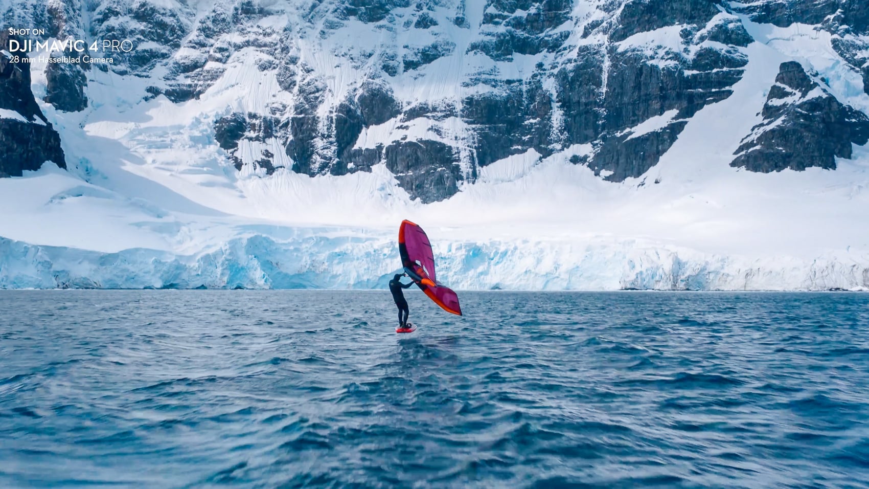 Wing foiler skimming cold open water below a towering snow-covered cliff.