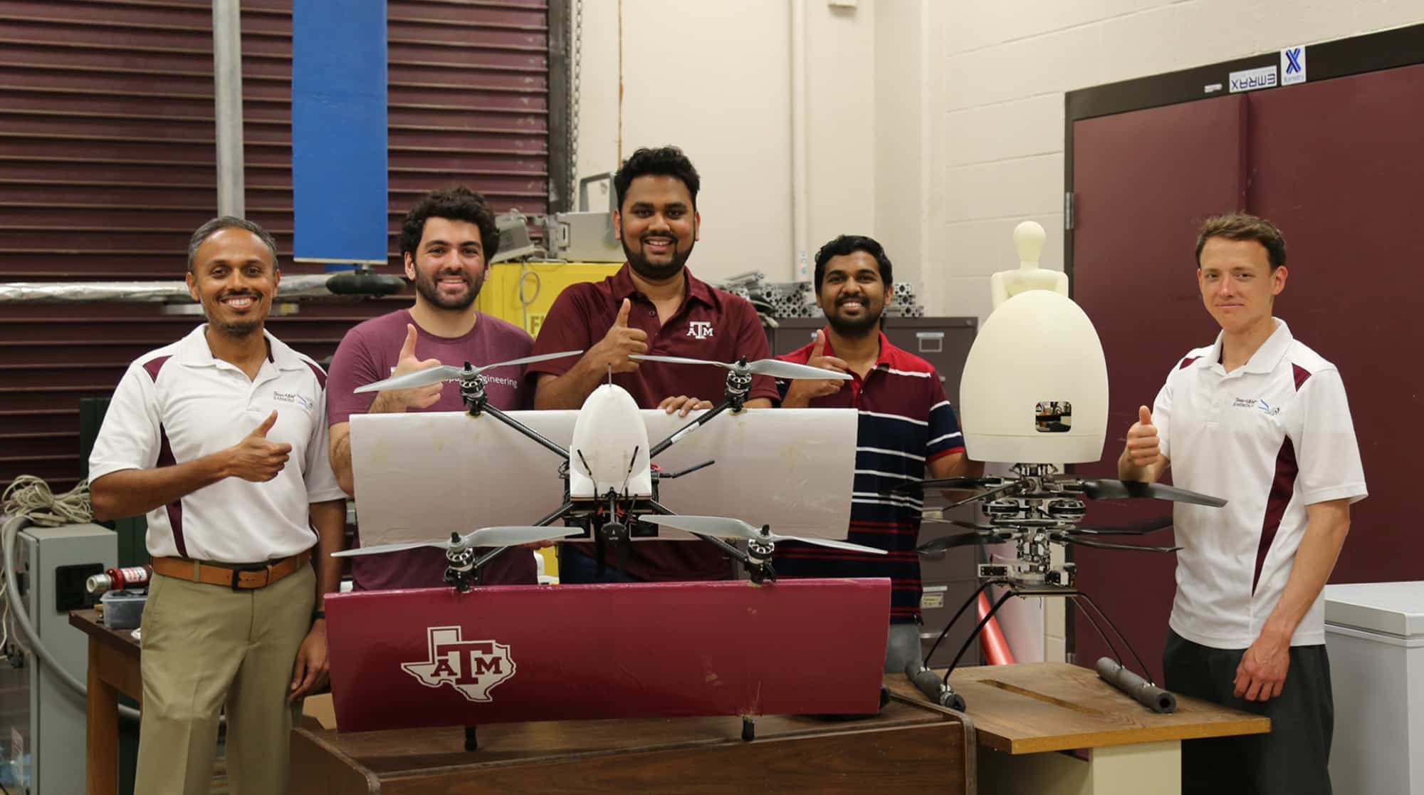 Five students in matching polos pose with two fixed-wing research drones in a lab.
