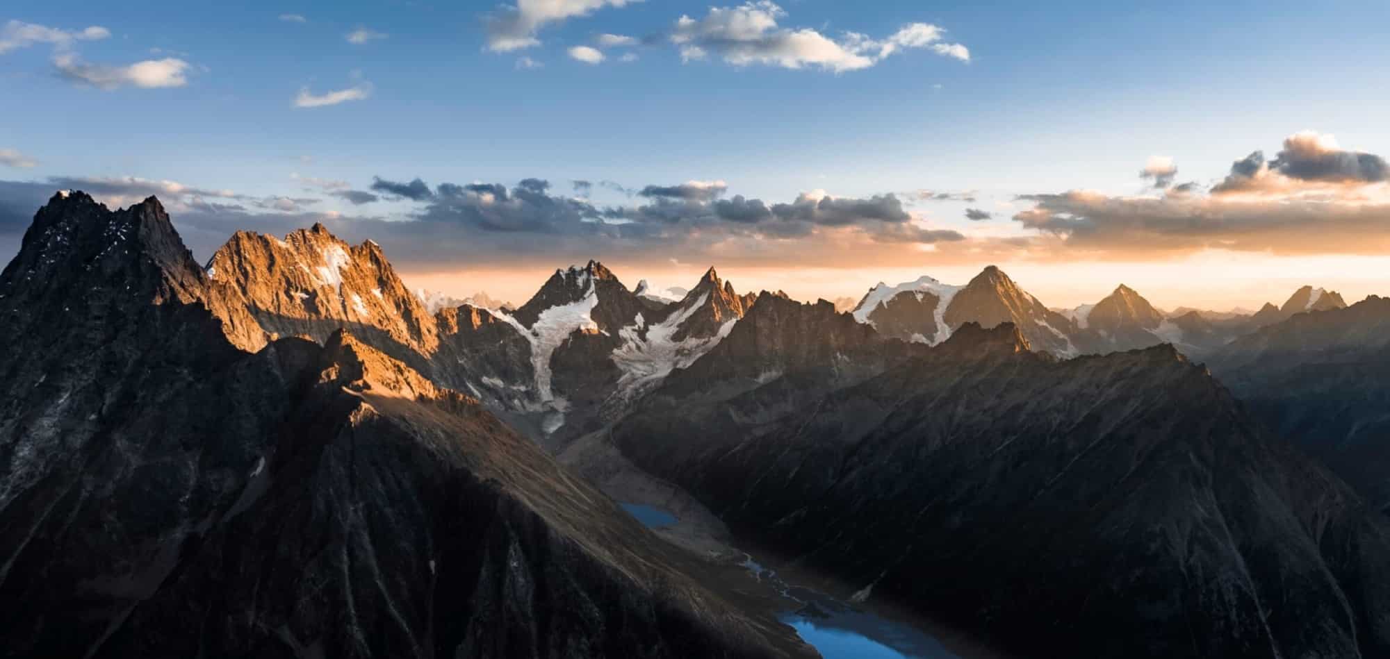 Aerial photo of jagged mountain peaks glowing at sunset above a small lake.