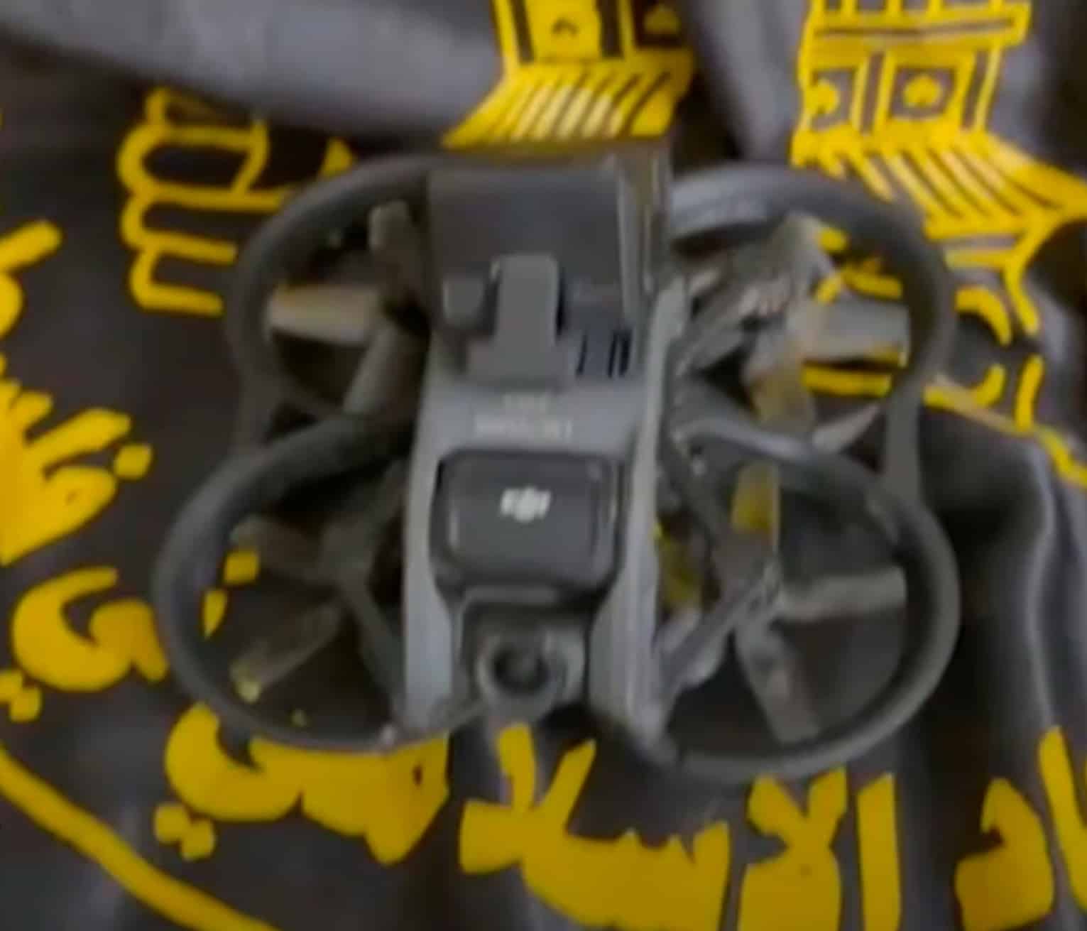 A captured FPV cinewhoop drone displayed on a black flag with yellow Arabic script.