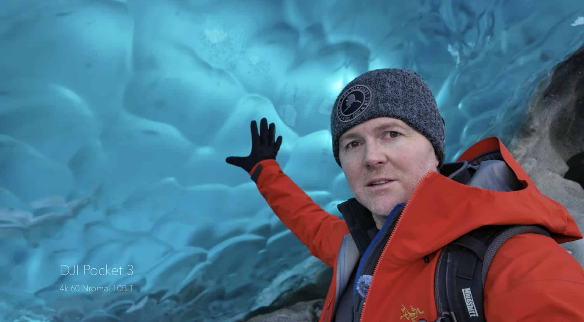 Man in a red jacket waving inside a blue glacier ice cave.