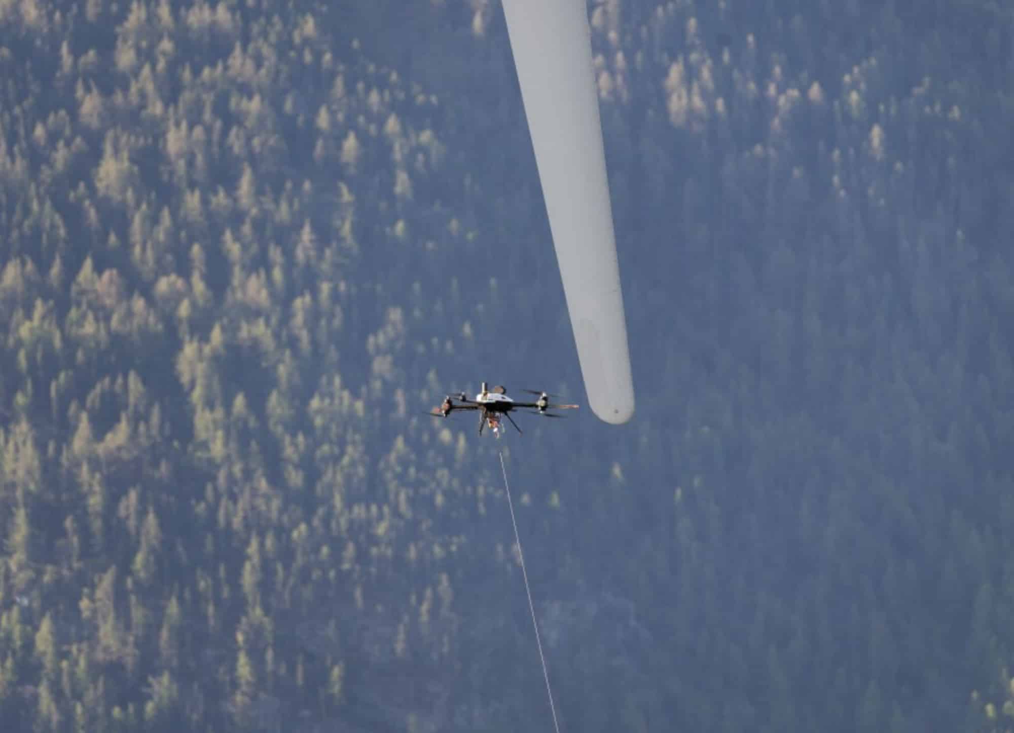 A tethered inspection drone hovers beside a wind turbine blade above dense forest.