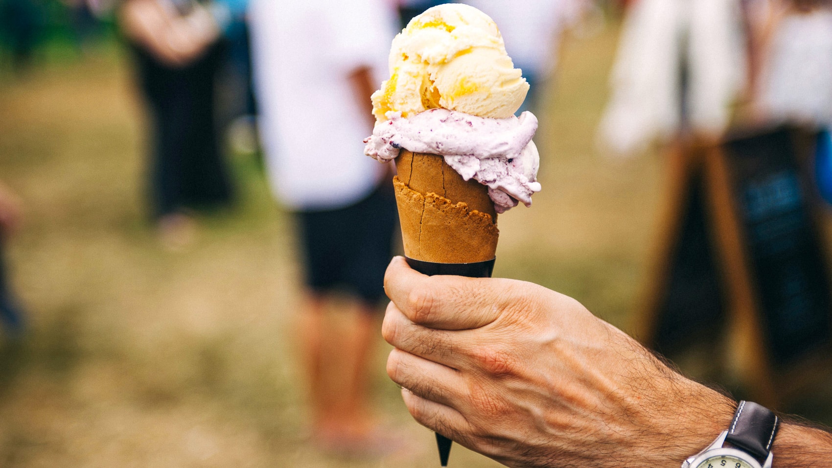 Hand holds a two-scoop ice cream cone in front of a blurred crowd.