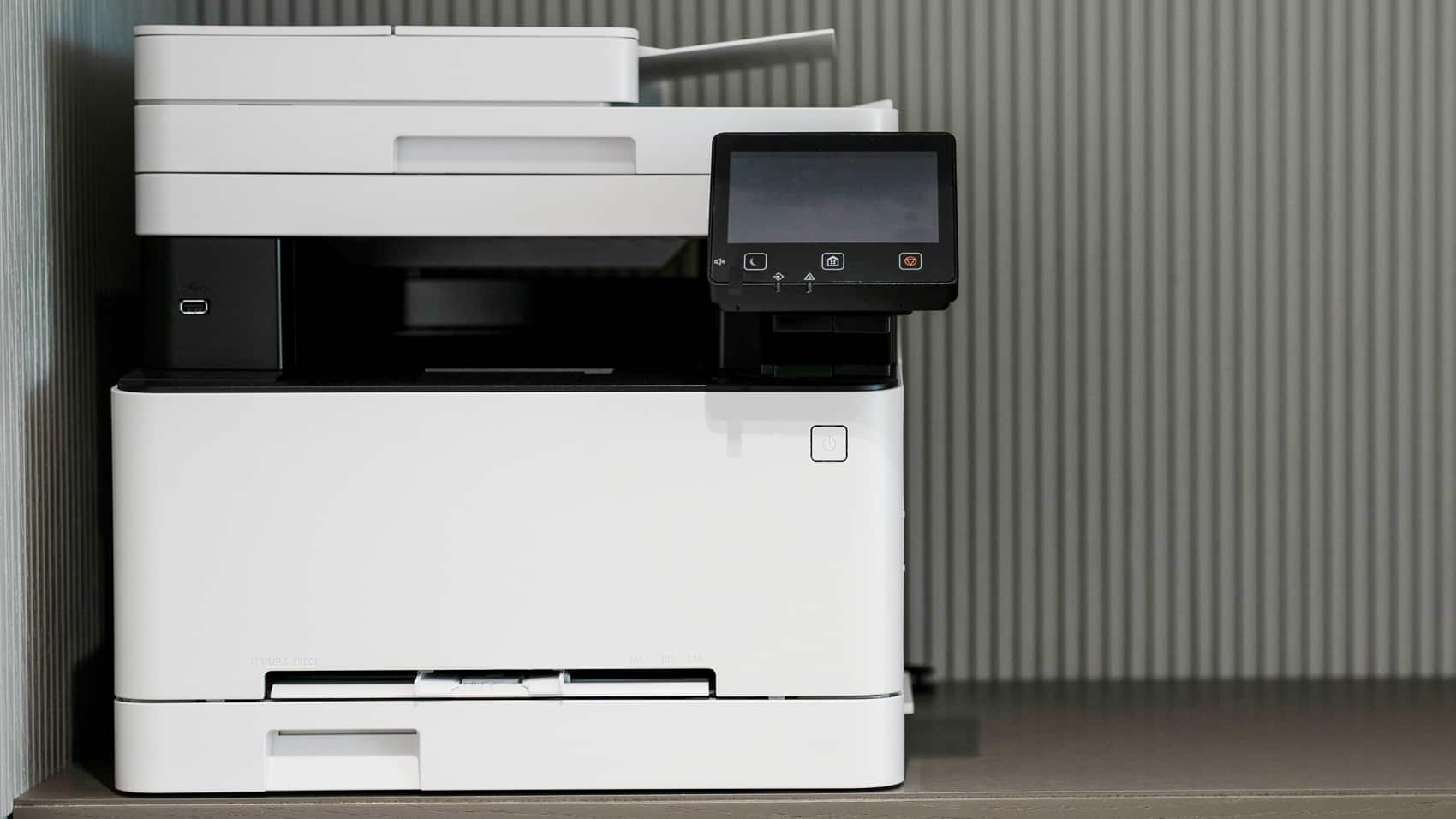 A white office multifunction printer stands against a plain wall.
