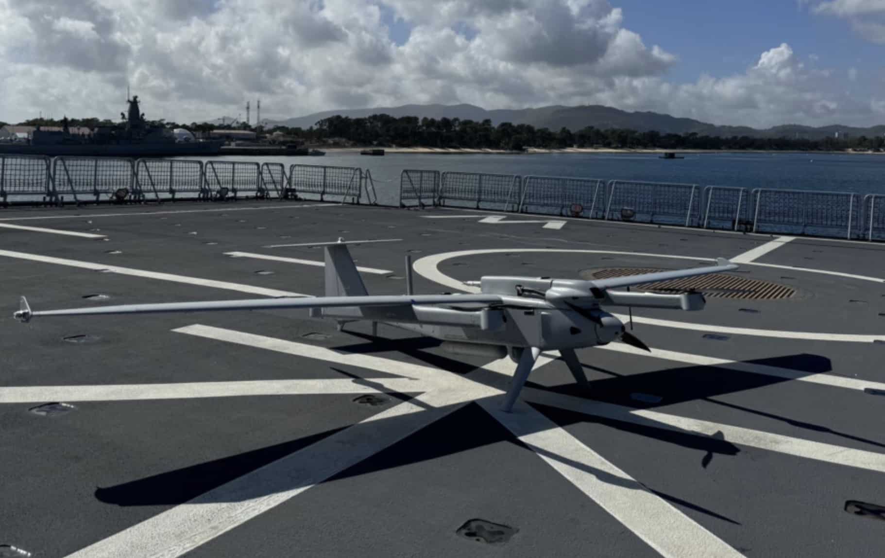 Gray fixed-wing VTOL drone parked on a warship flight deck beside a harbor.