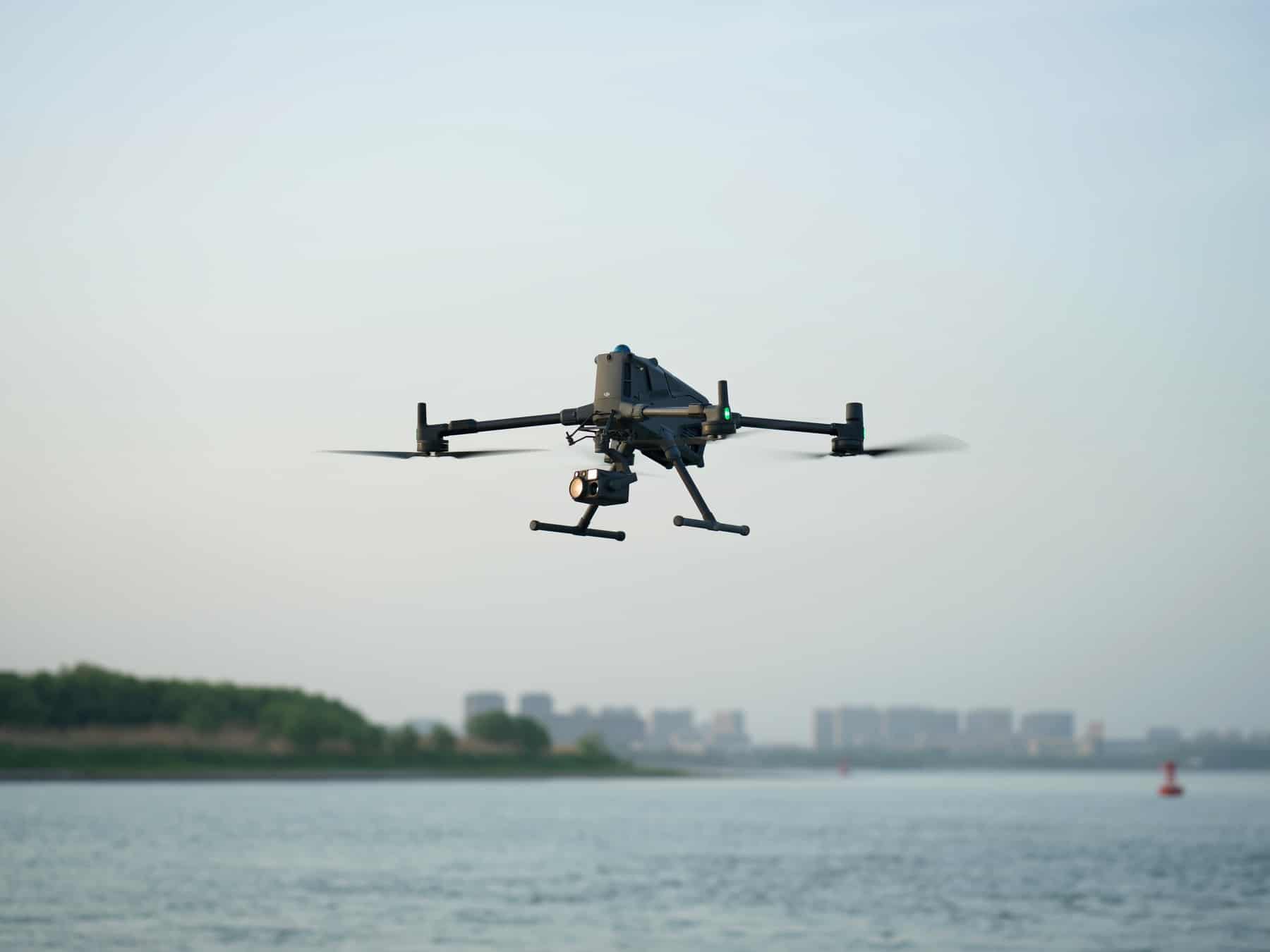 The Matrice 400 hovers over a wide river with a hazy city skyline behind.