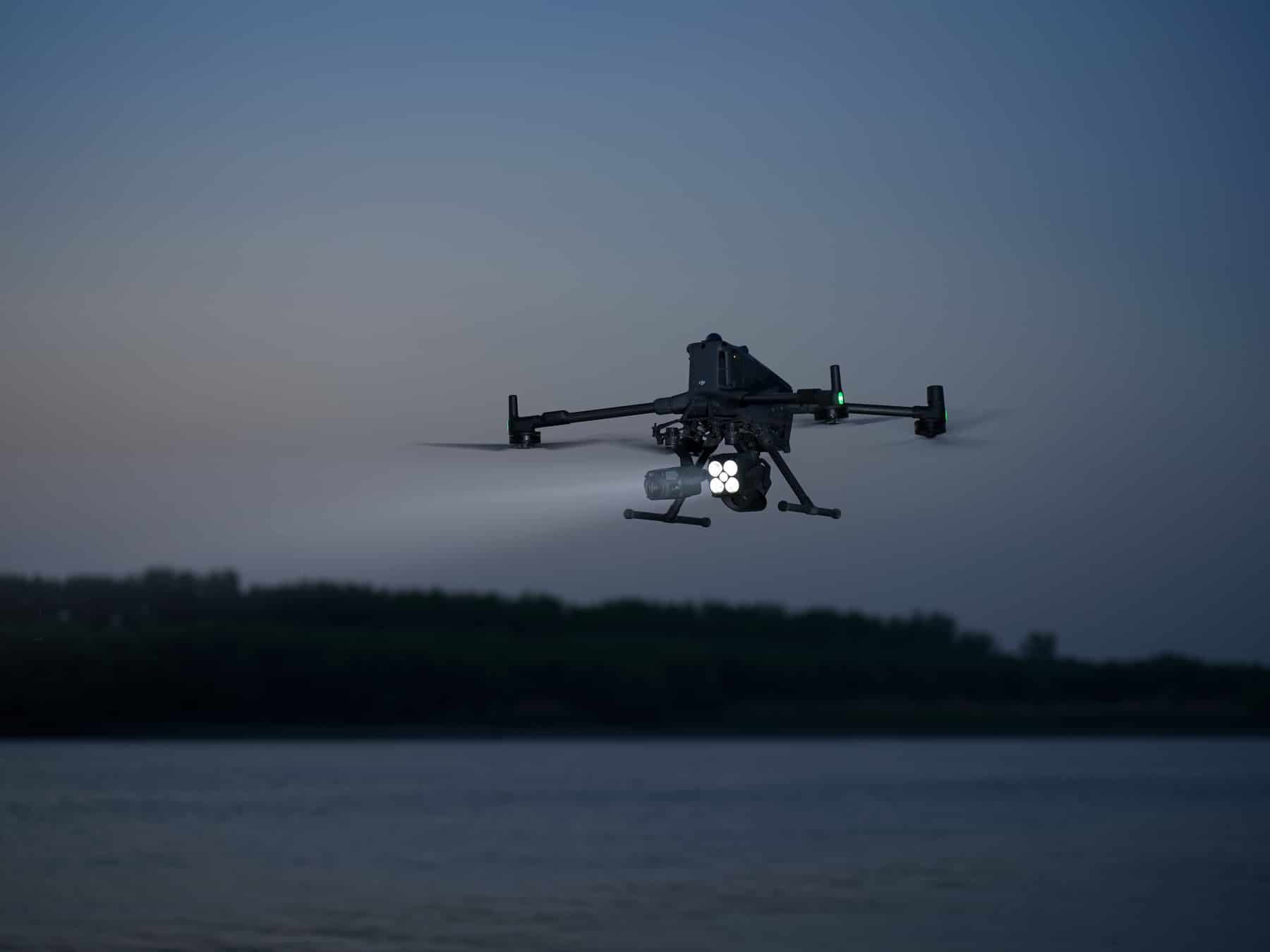 Enterprise quadcopter hovers above dark water at dusk with its payload lights on.