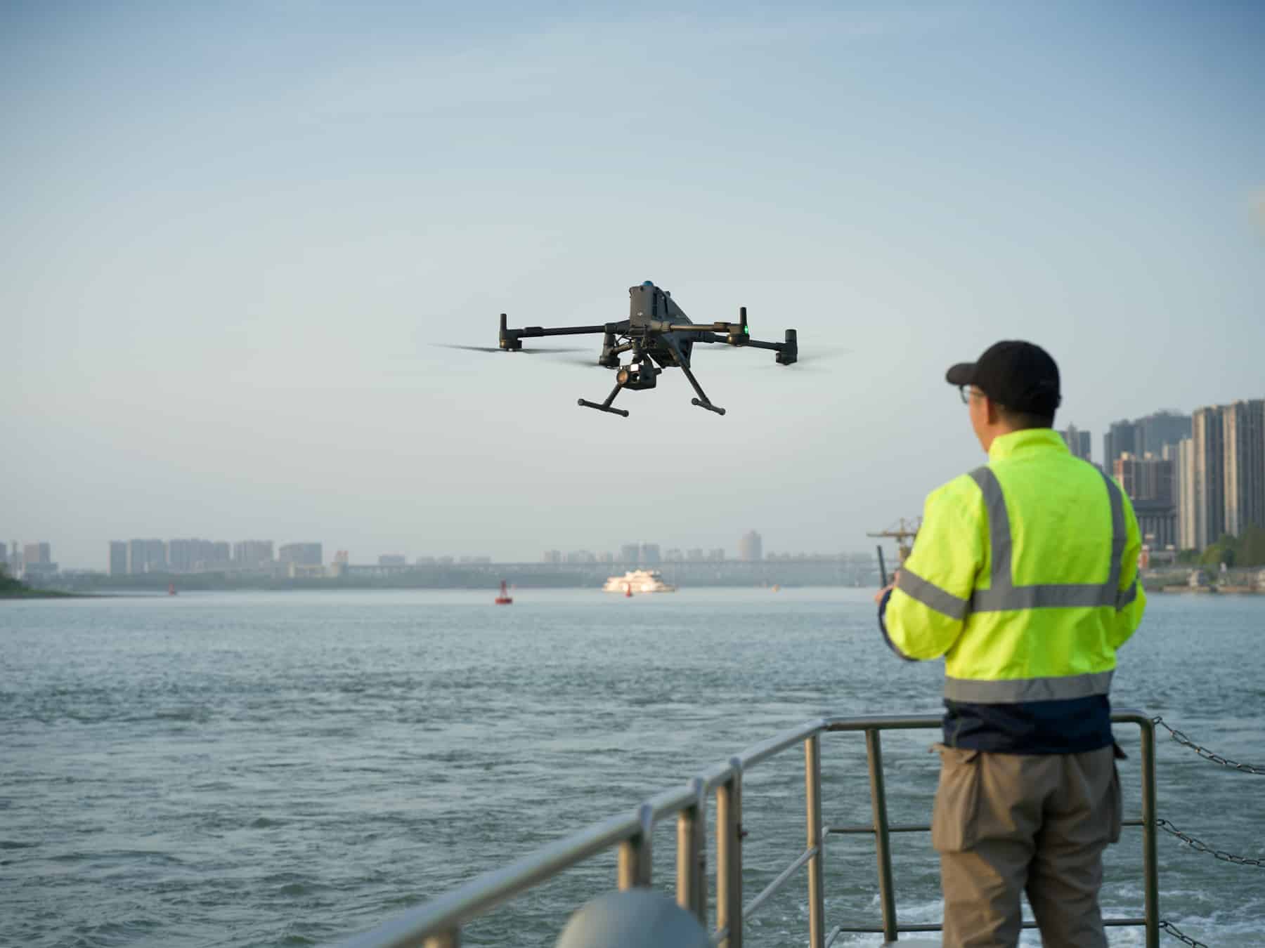 Crew member in a high-visibility vest watches the drone from a boat railing.