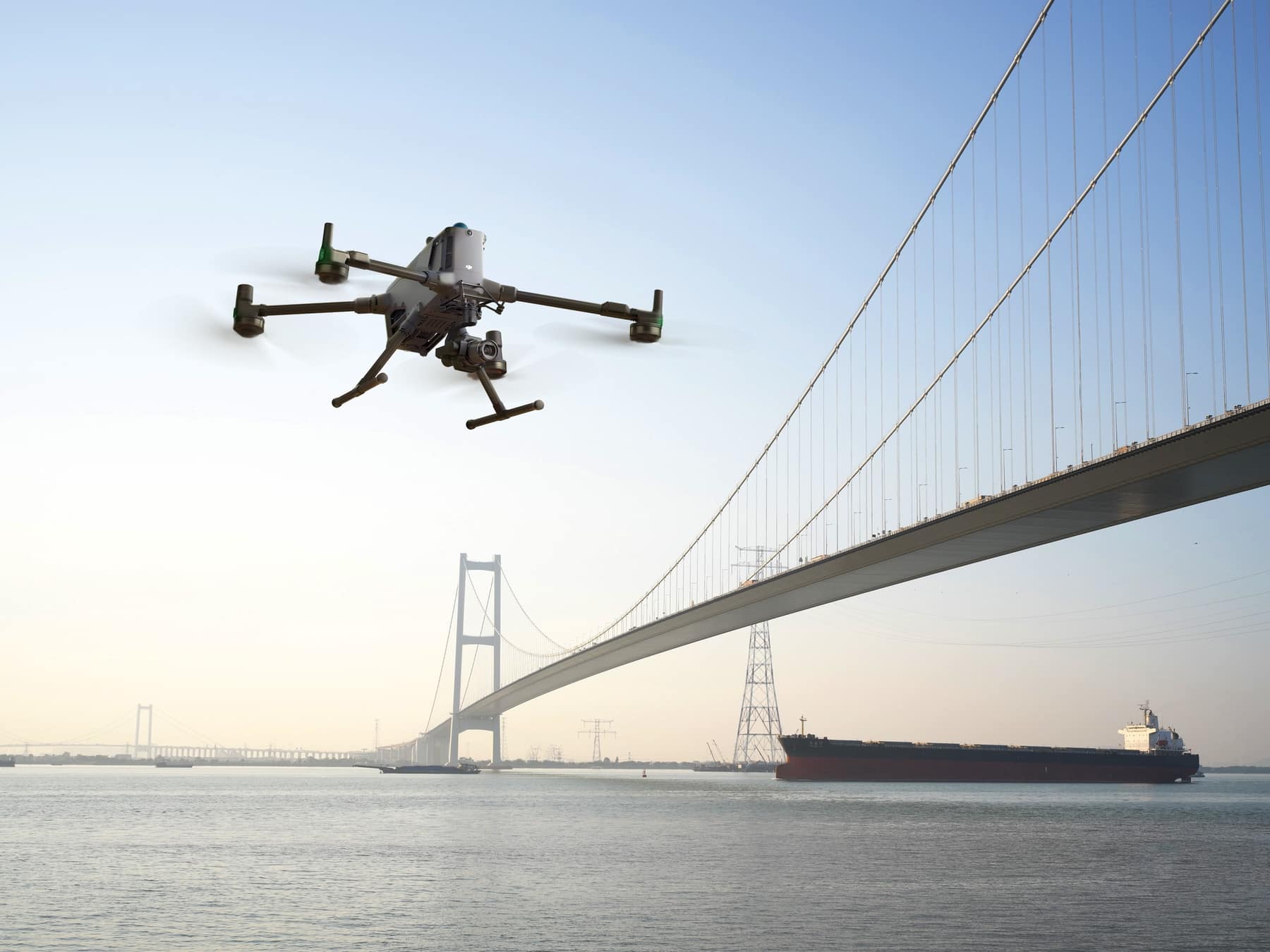 An enterprise quadcopter hovers above a bay spanned by a long suspension bridge.
