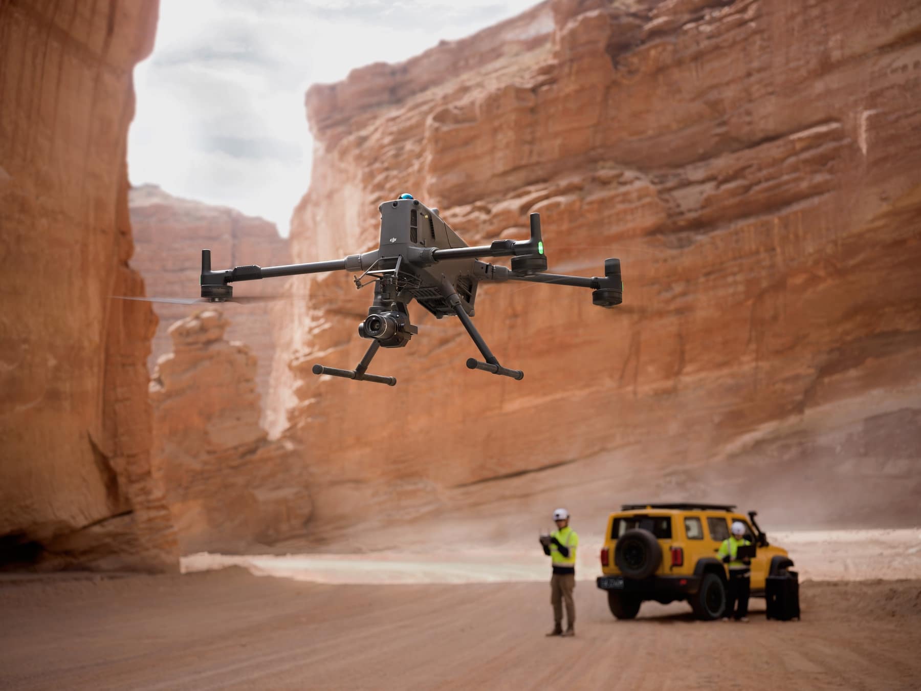 Drone hovers in a narrow gorge above a yellow off-road vehicle and crew.