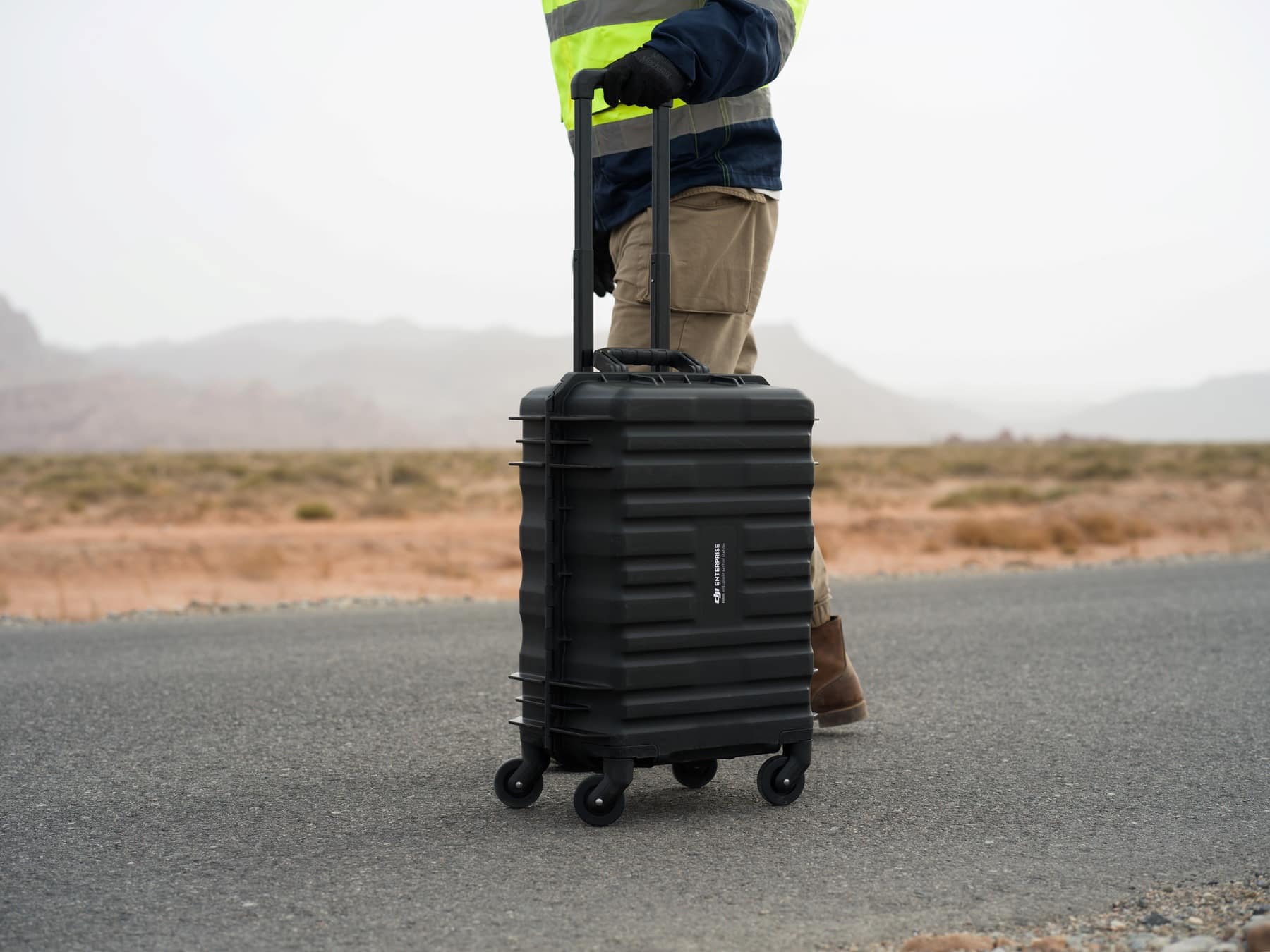 Worker in a high-visibility vest wheels a black rolling case along a road.