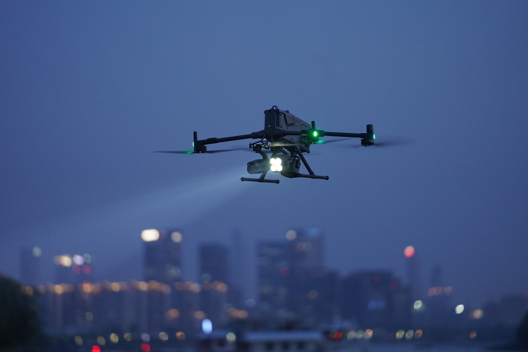 At night the drone shines a spotlight beam over a lit waterfront city.