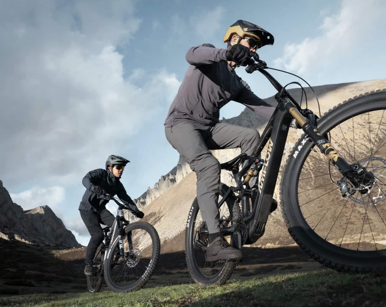 Two riders climb a dirt hillside on full-suspension electric mountain bikes.