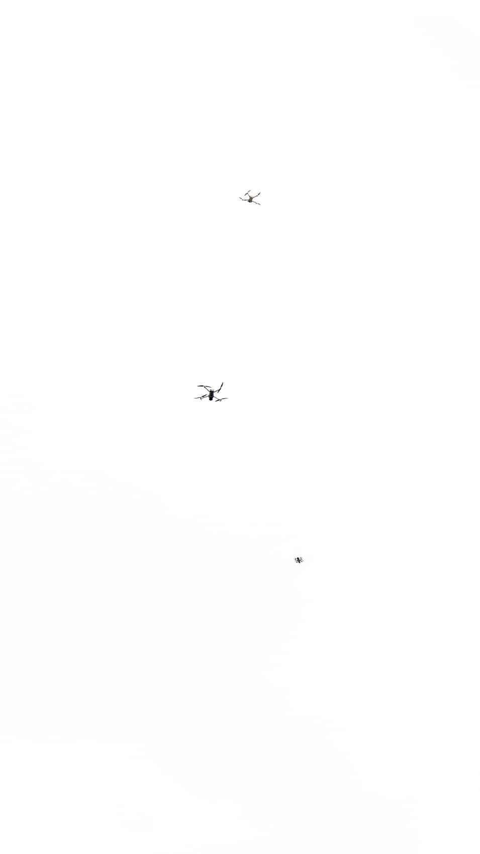 Three small drones fly at different heights against a featureless white overcast sky.