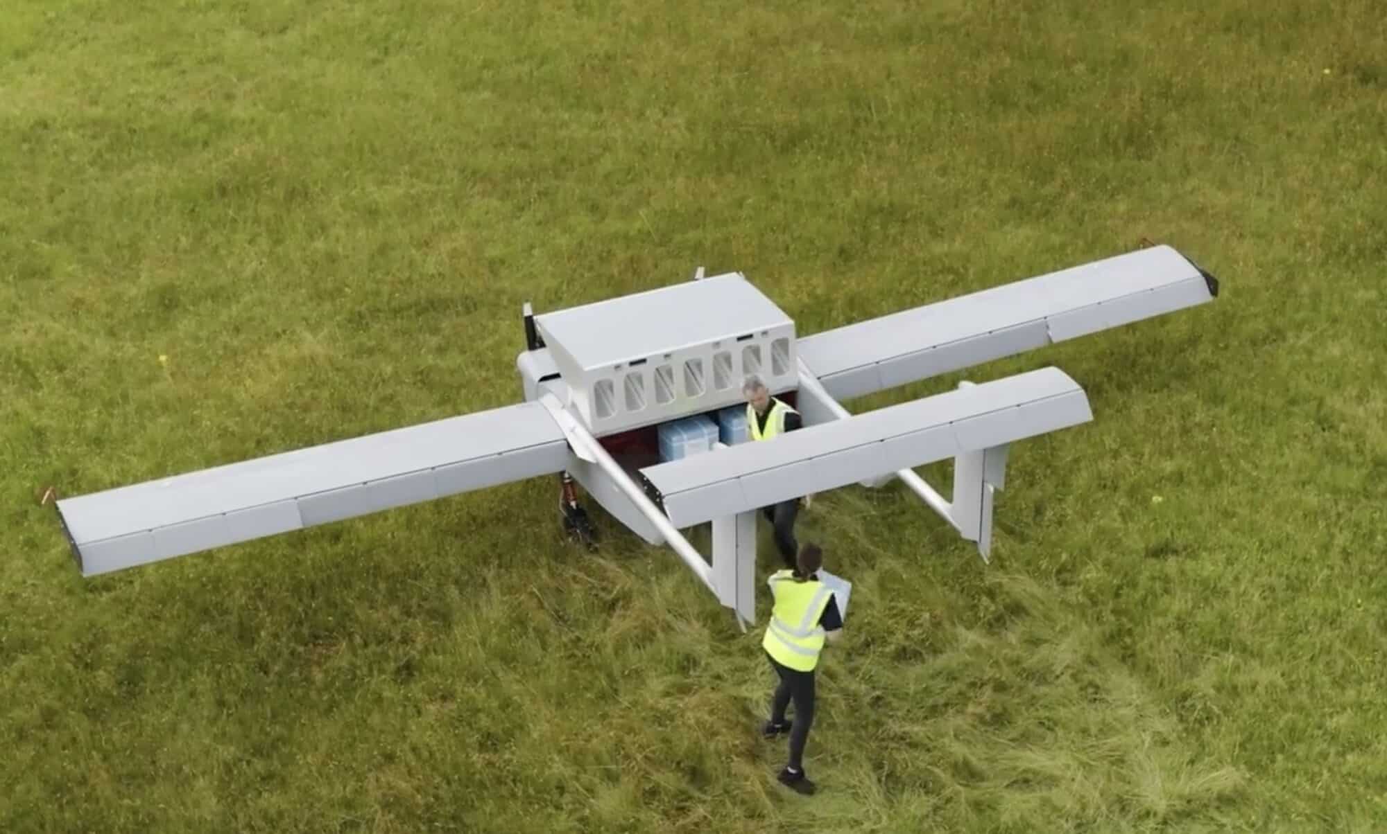 Large white box-wing cargo drone parked on grass beside a worker in hi-vis.