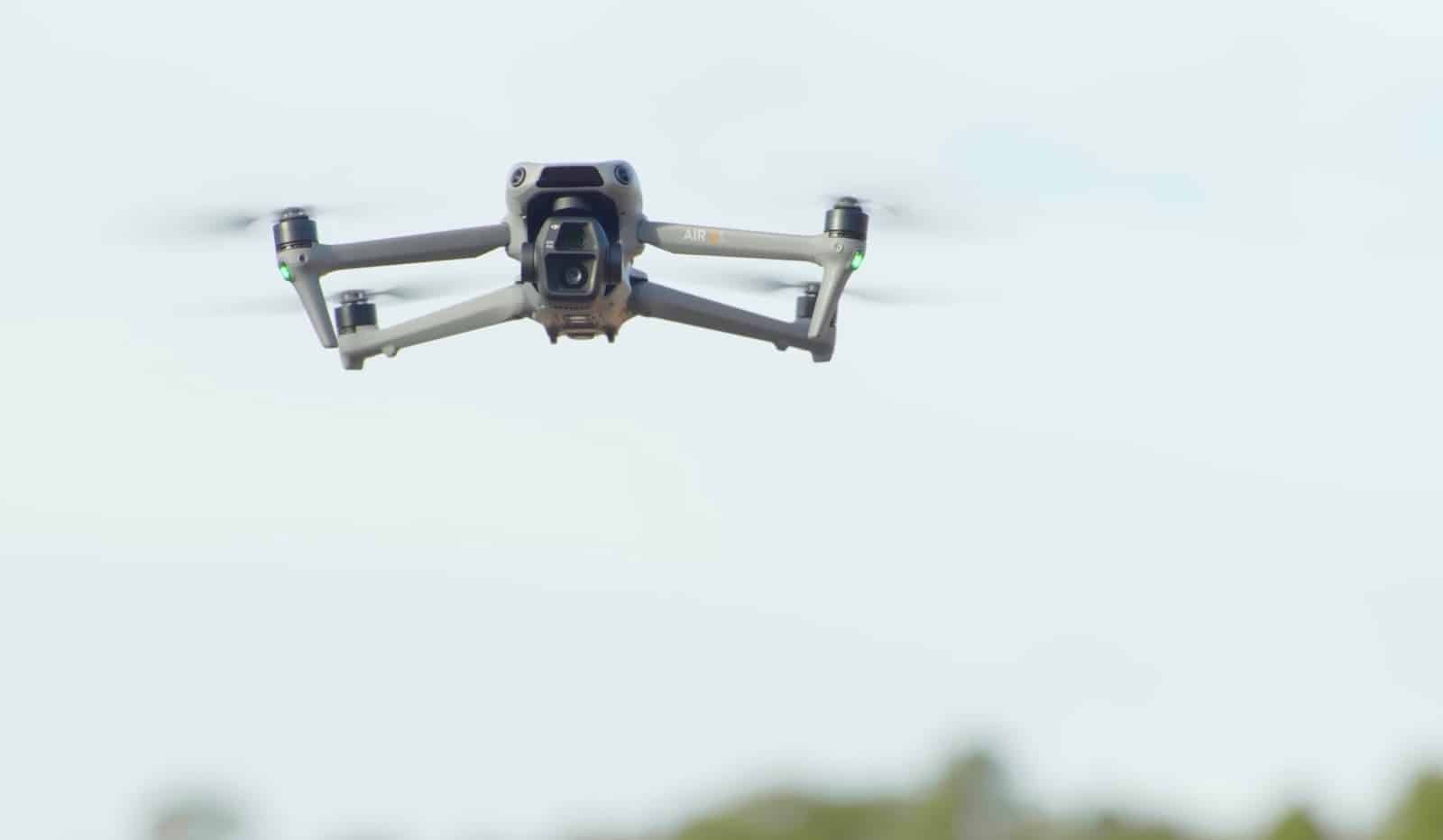A grey folding quadcopter hovers against a washed-out pale sky.