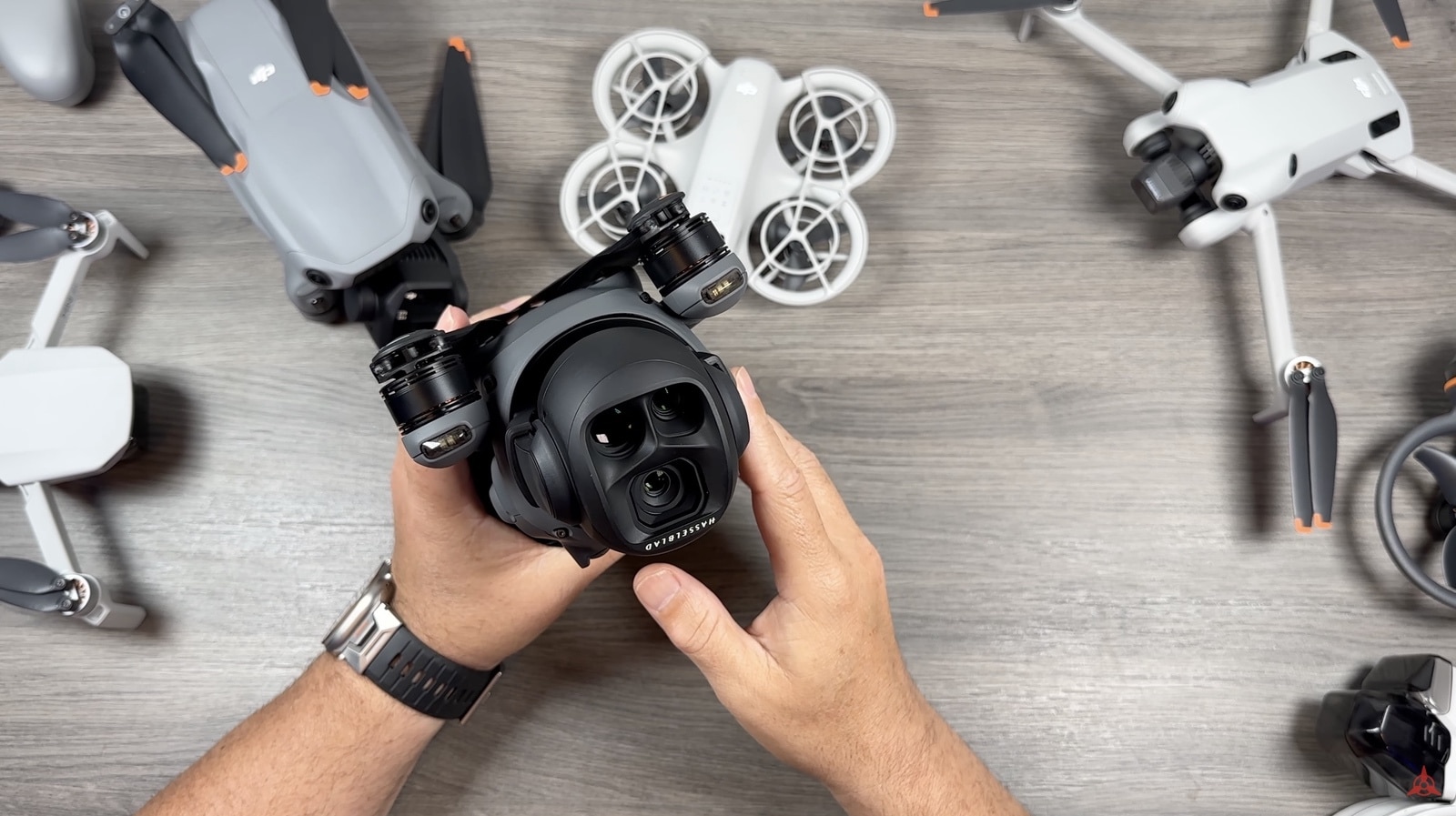 Hands lift a Mavic 4 Pro by its triple-camera gimbal among other DJI drones.