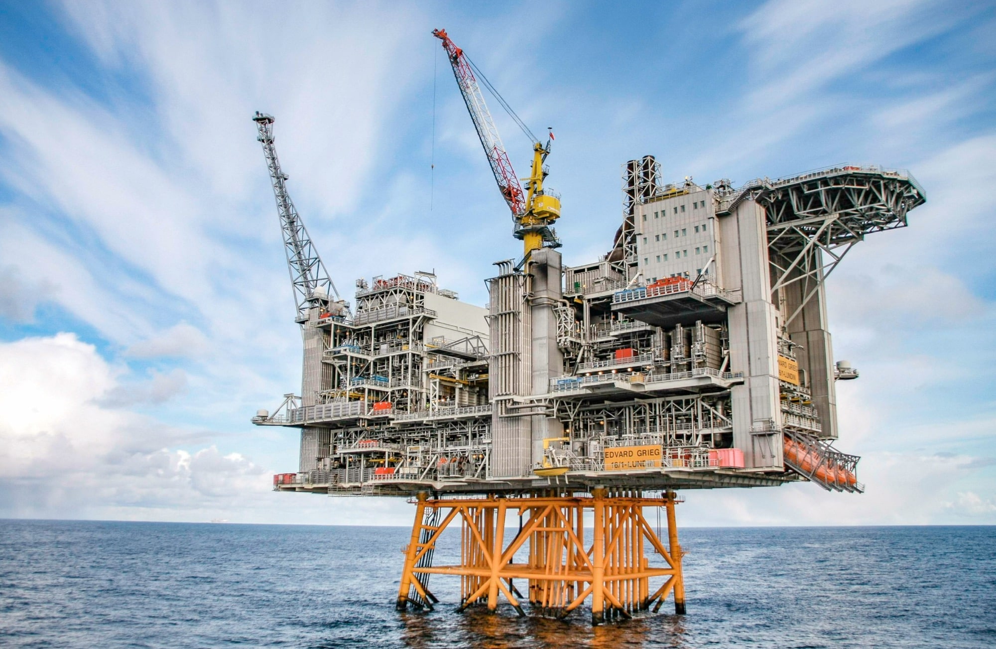 An offshore oil production platform stands over calm blue water under scattered cloud.