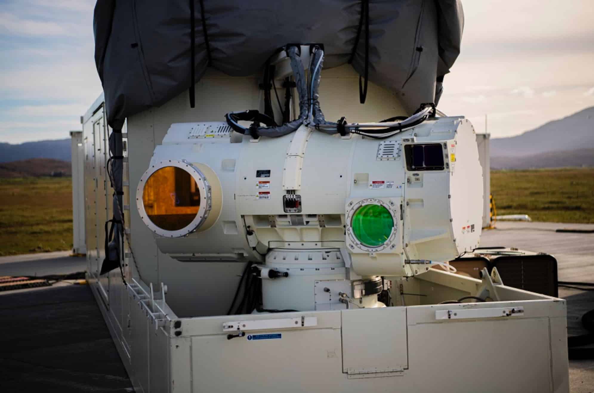 A container-mounted laser turret with orange and green optical apertures faces open moorland.