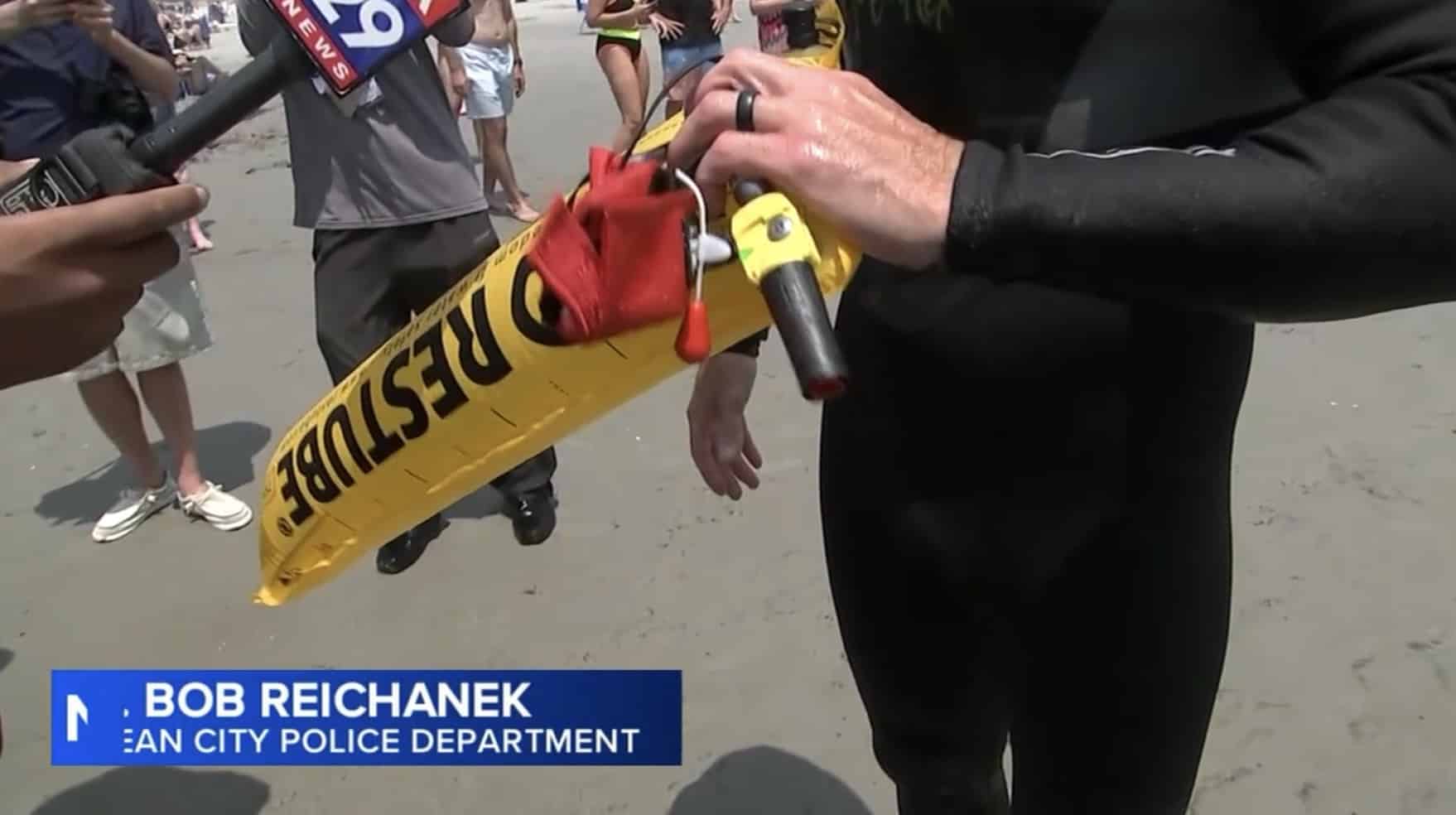 Wetsuited hands holding a yellow Restube flotation device as news microphones crowd in.