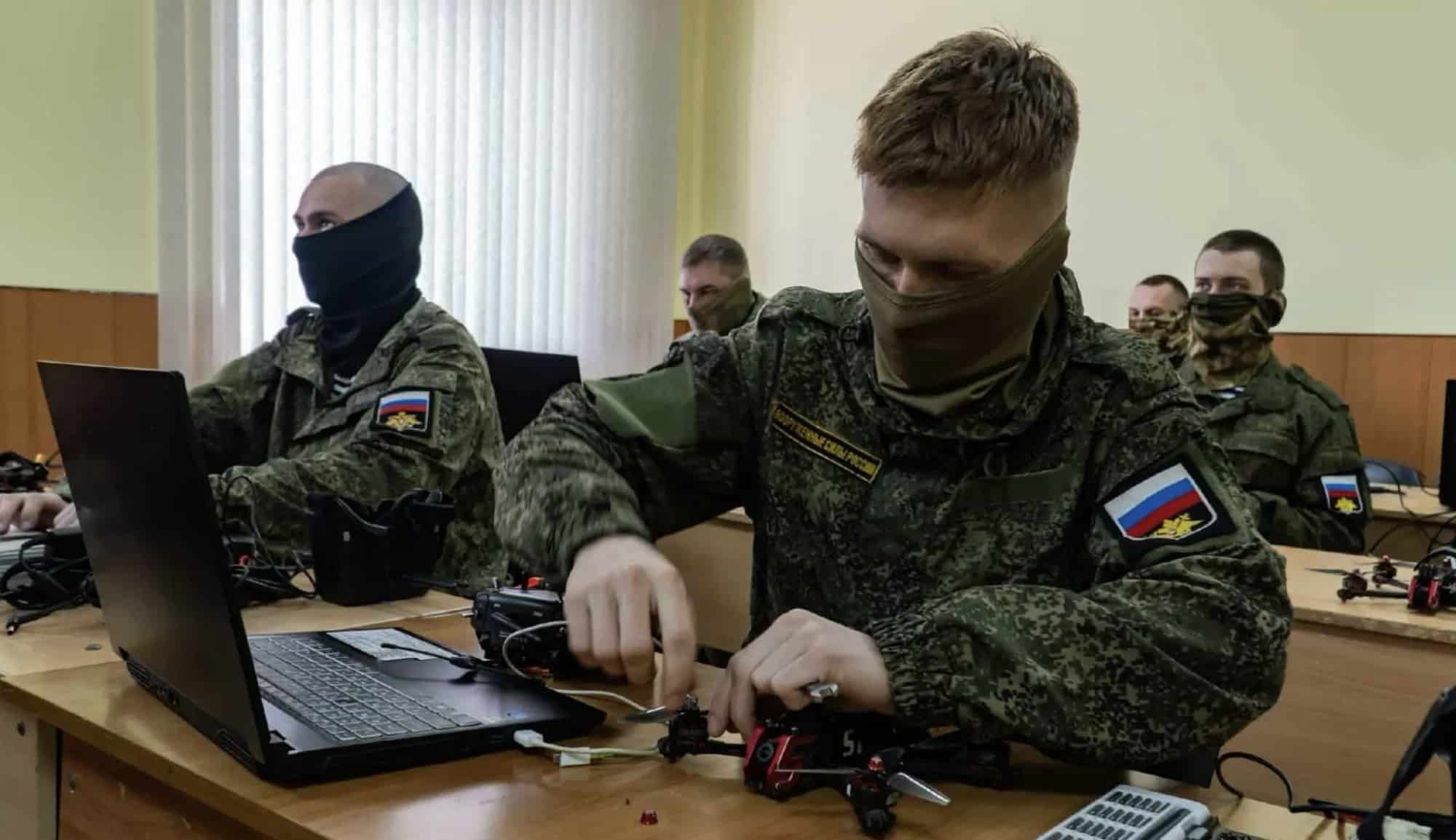 Soldiers with Russian flag patches assemble small drone parts at classroom desks.