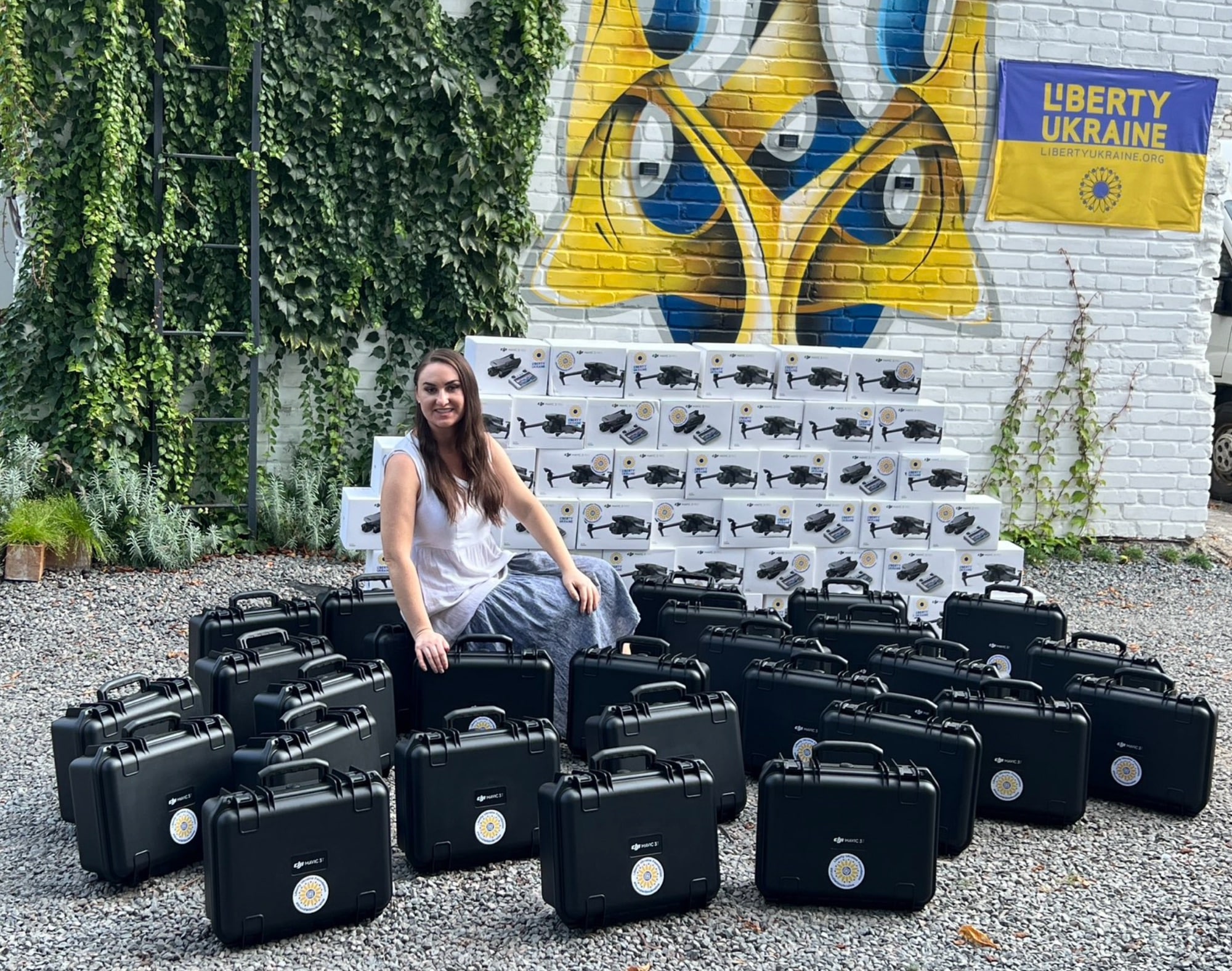 Woman seated among dozens of stacked drone cases before a mural and a Ukraine flag.