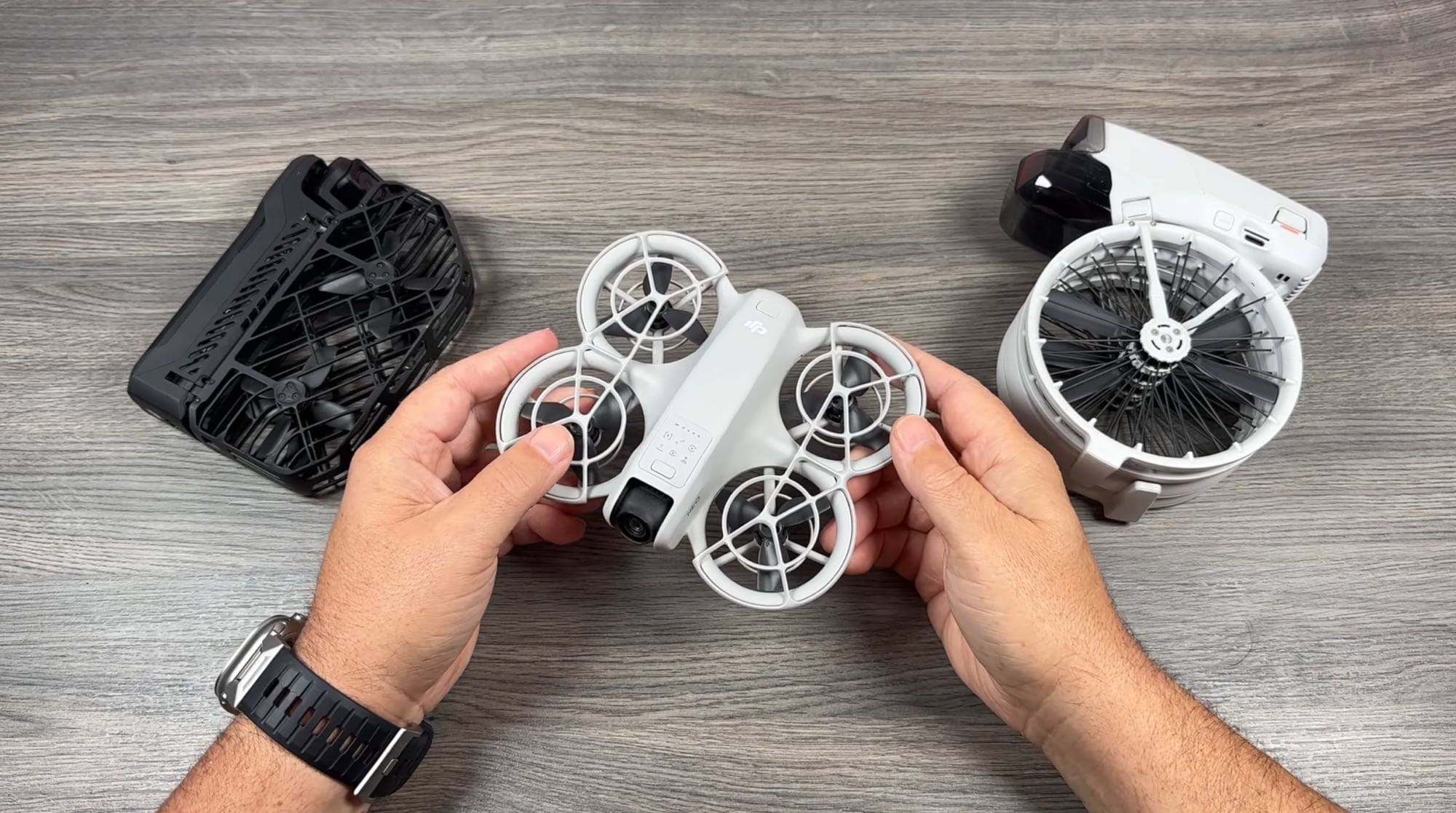 Hands lift the white DJI Neo from between a HoverAir X1 and DJI Flip.