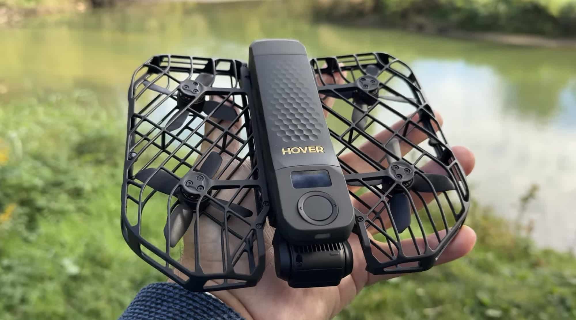 A hand holds the black HoverAir X1 Pro Max beside a green riverbank.