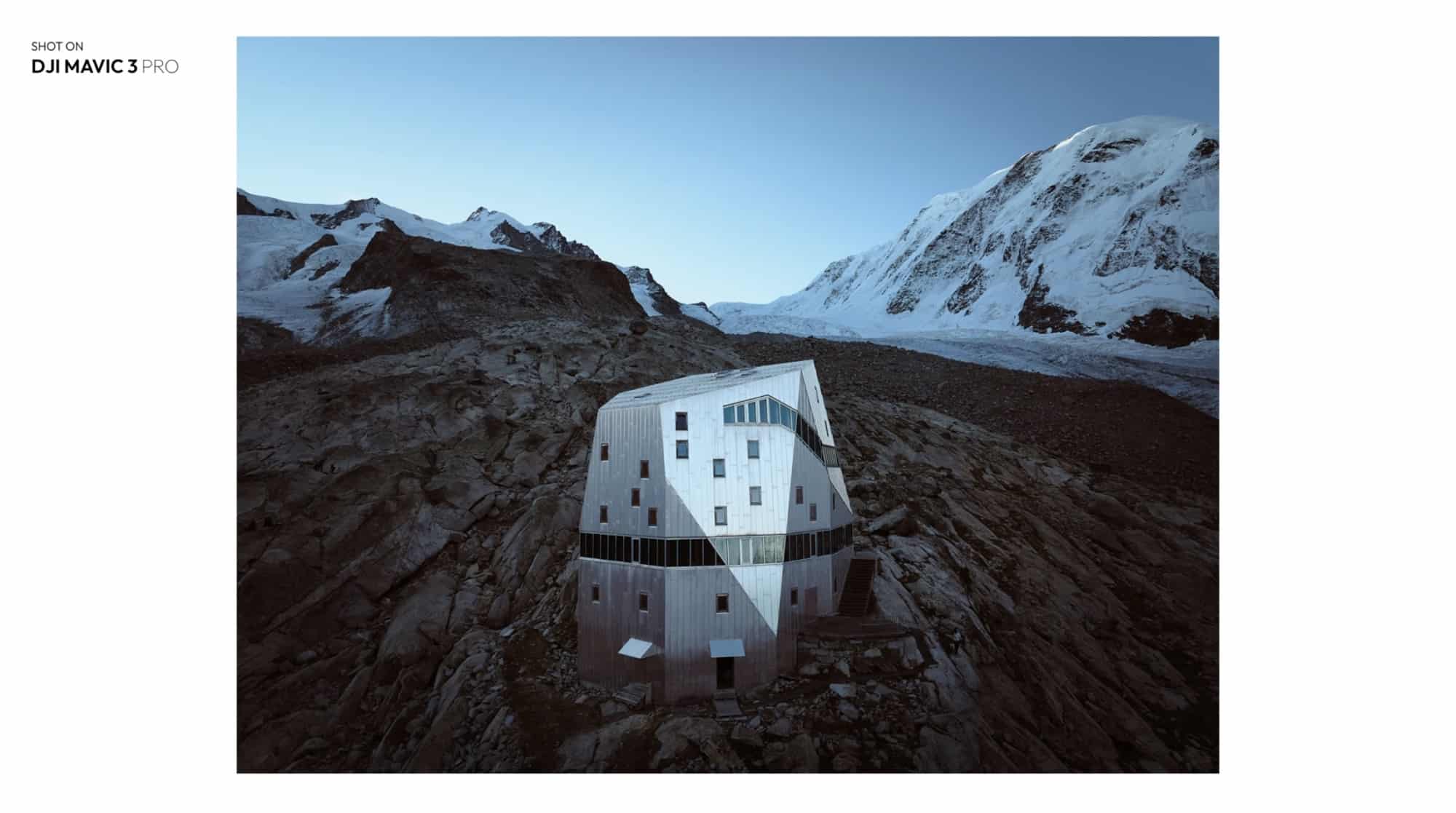 Angular white alpine hut on a rocky ridge below snowy peaks, credited to the Mavic 3 Pro.