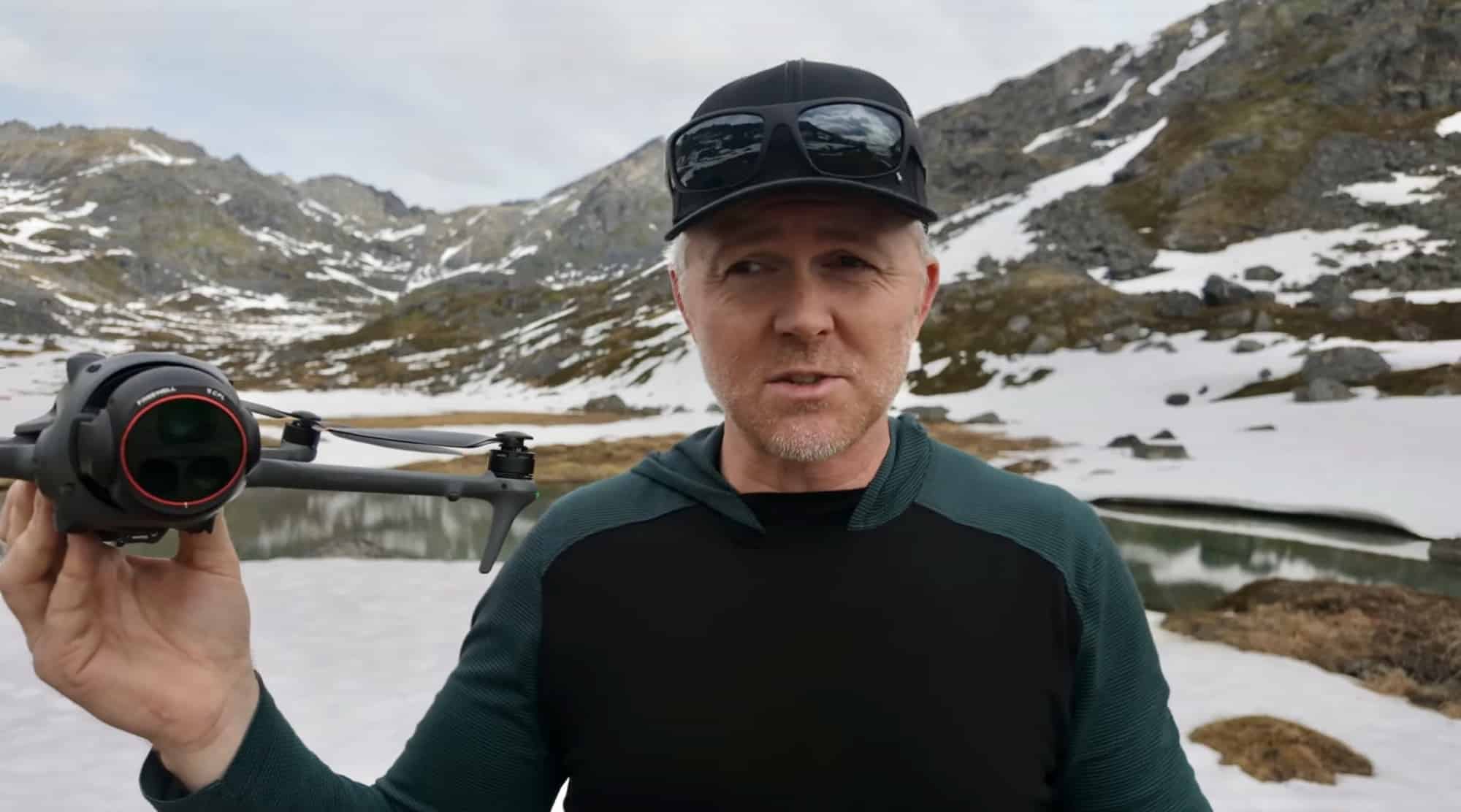 The reviewer holds the drone toward the camera in a snow-lined alpine valley.