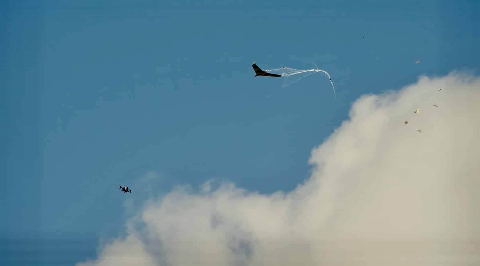 A tiny drone trails a deployed net against bright sky and scattered cloud.