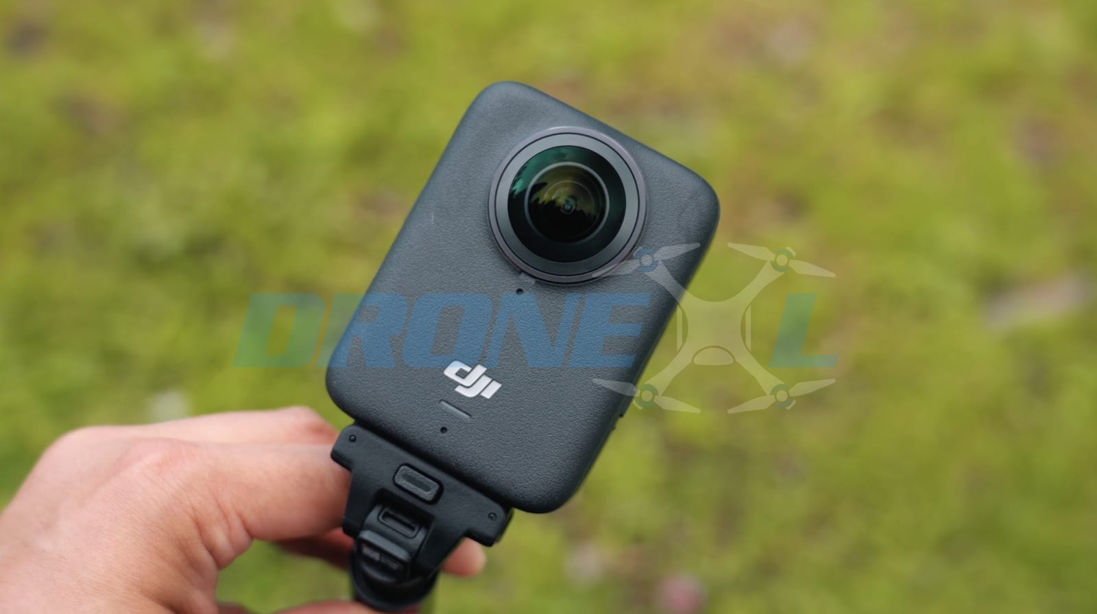 Hand holding the camera over green grass, showing its rear lens and mount.
