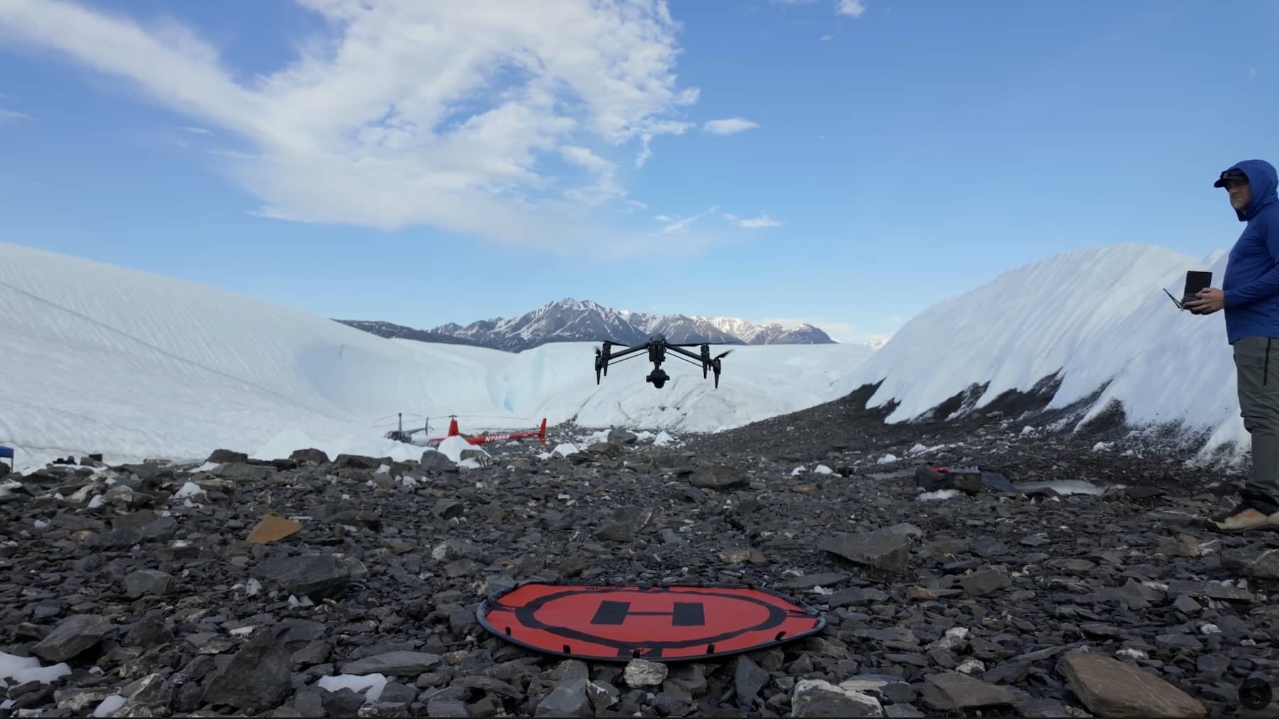 Pilot works beside a red landing pad on a glacier moraine as a drone lifts off.