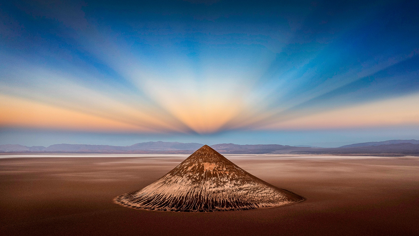 Aerial of a lone cone-shaped hill on a desert plain at sunset.
