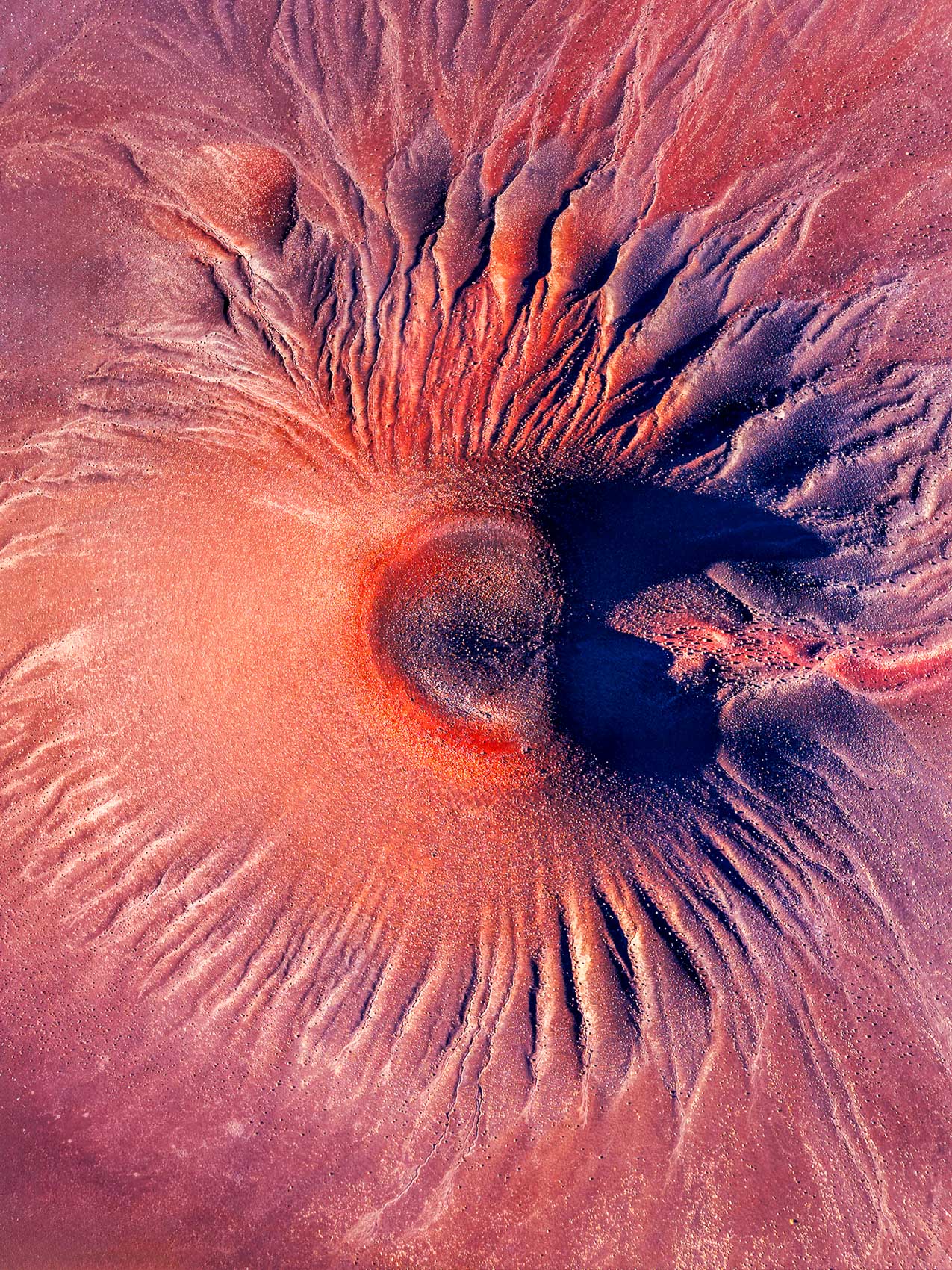 Overhead abstract of a pink crater ringed by radiating sand ridges.