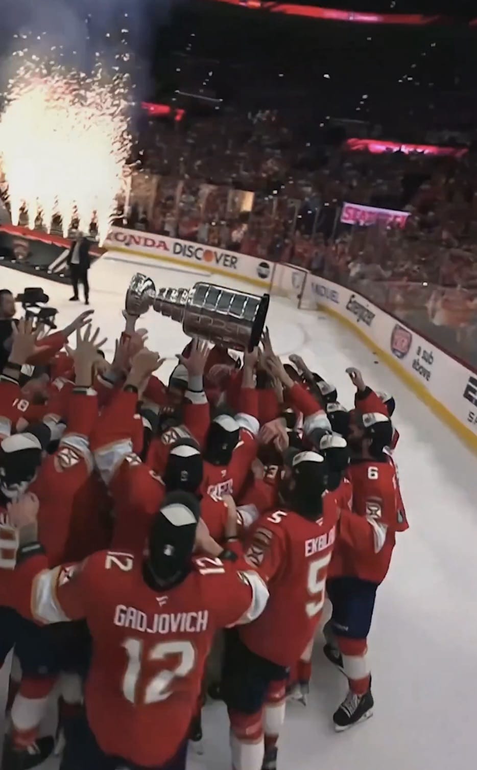Drone view over hockey players celebrating on the ice amid arena pyrotechnics.