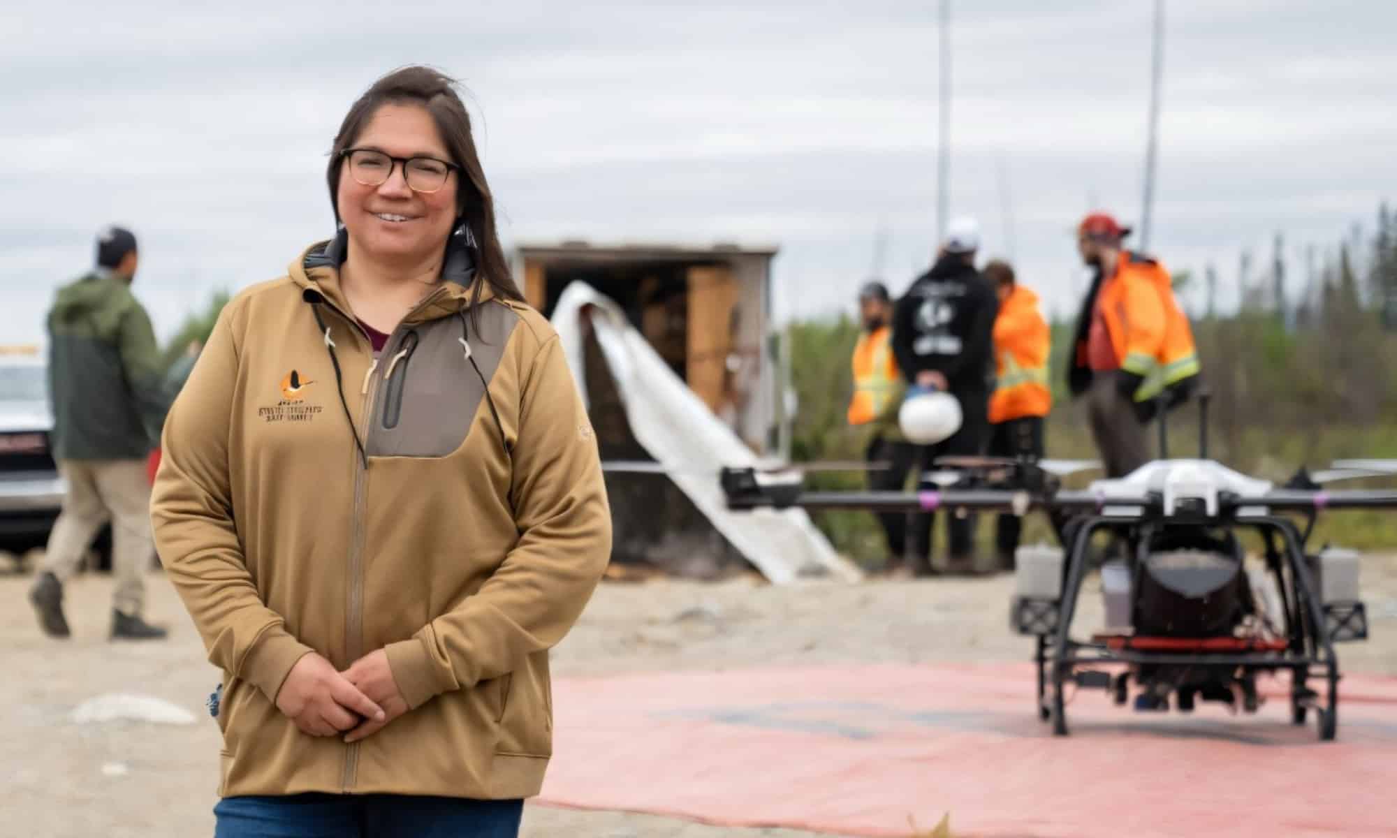 Woman in a tan jacket smiling in front of a seeding drone and a crew of workers.