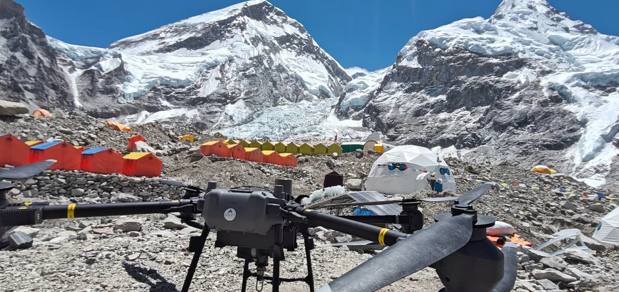 DJI FlyCart 30 parked on rocky moraine among orange tents below Himalayan peaks.