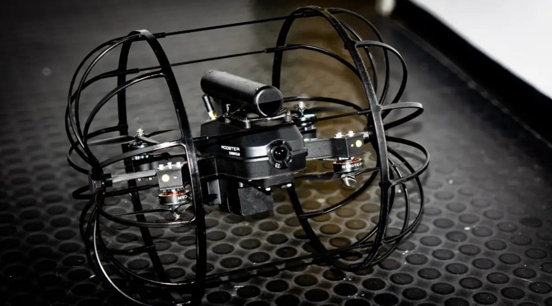 The caged Rooster drone resting on a dark rubber mat under studio lighting.