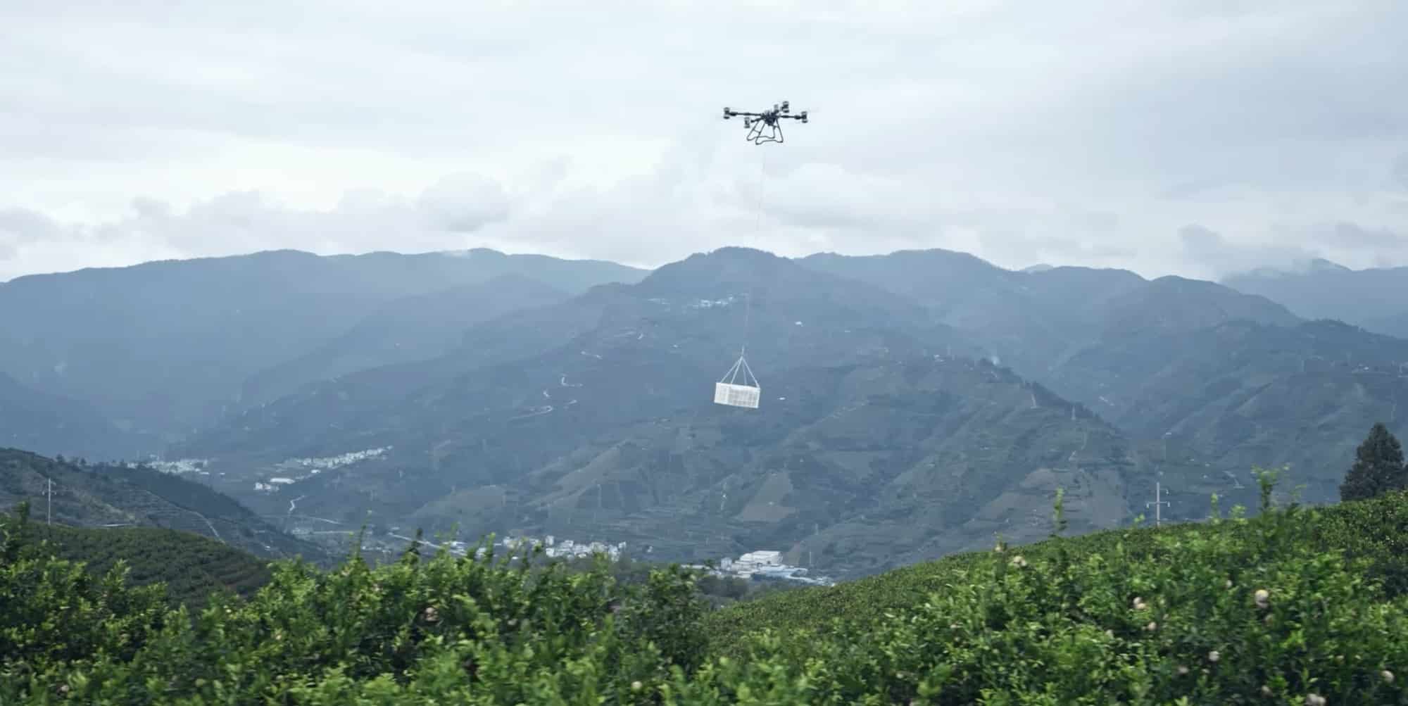 Agras drone carrying a slung load above terraced orchards in a mountain valley.