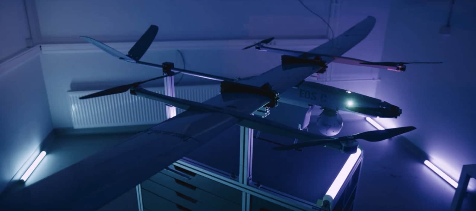 A quad-rotor VTOL fixed-wing drone rests on a stand in a blue-lit workshop.