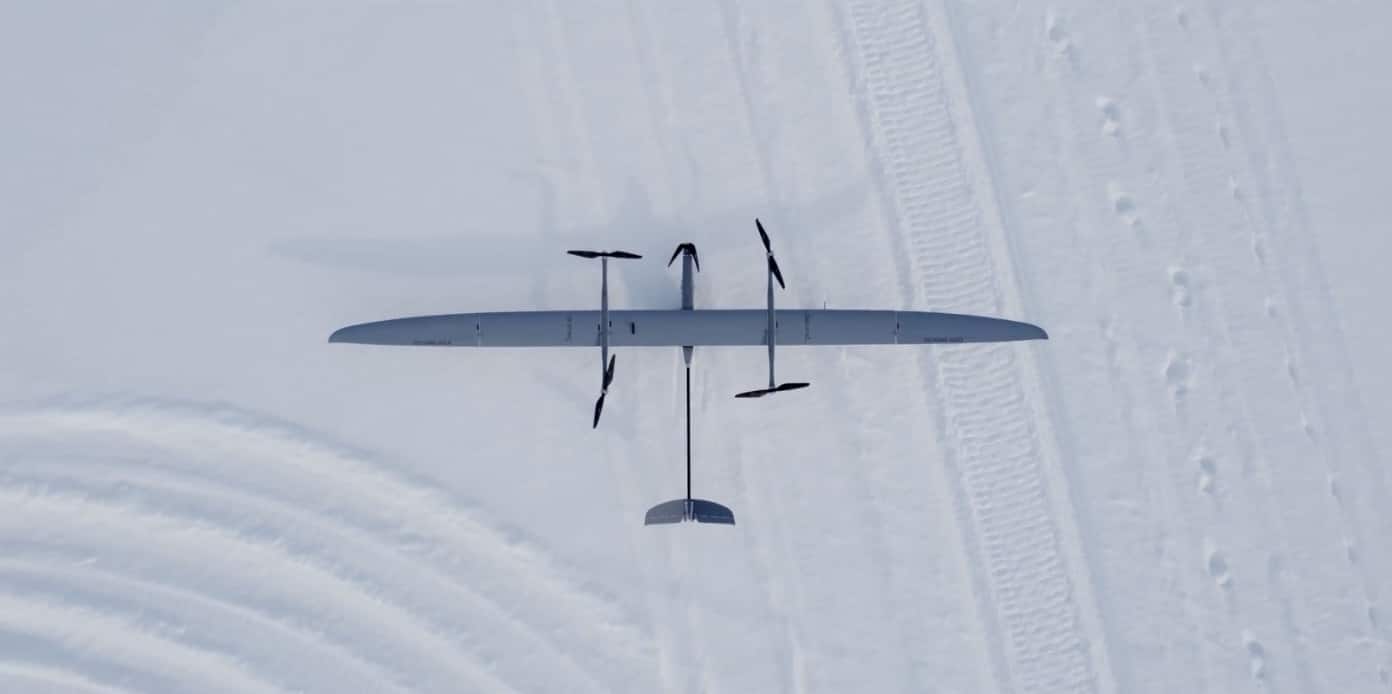 A fixed-wing surveillance drone banks above snow-covered ground crossed by vehicle tracks.