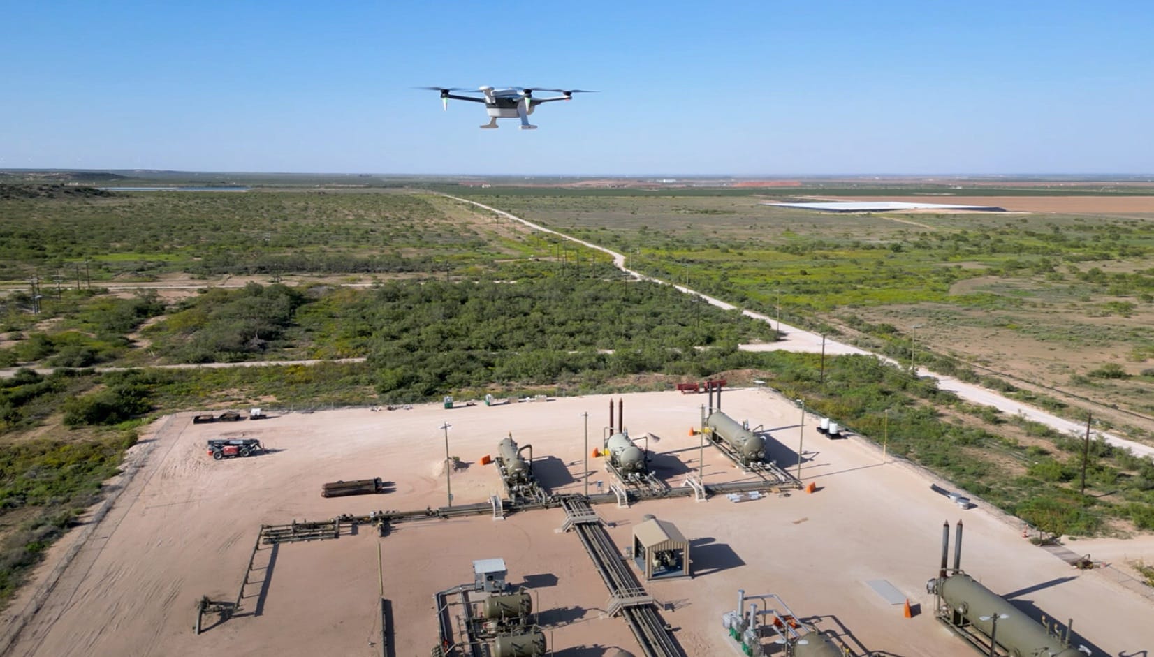 A quadcopter hovers above an oilfield wellpad ringed by pipework and scrub.
