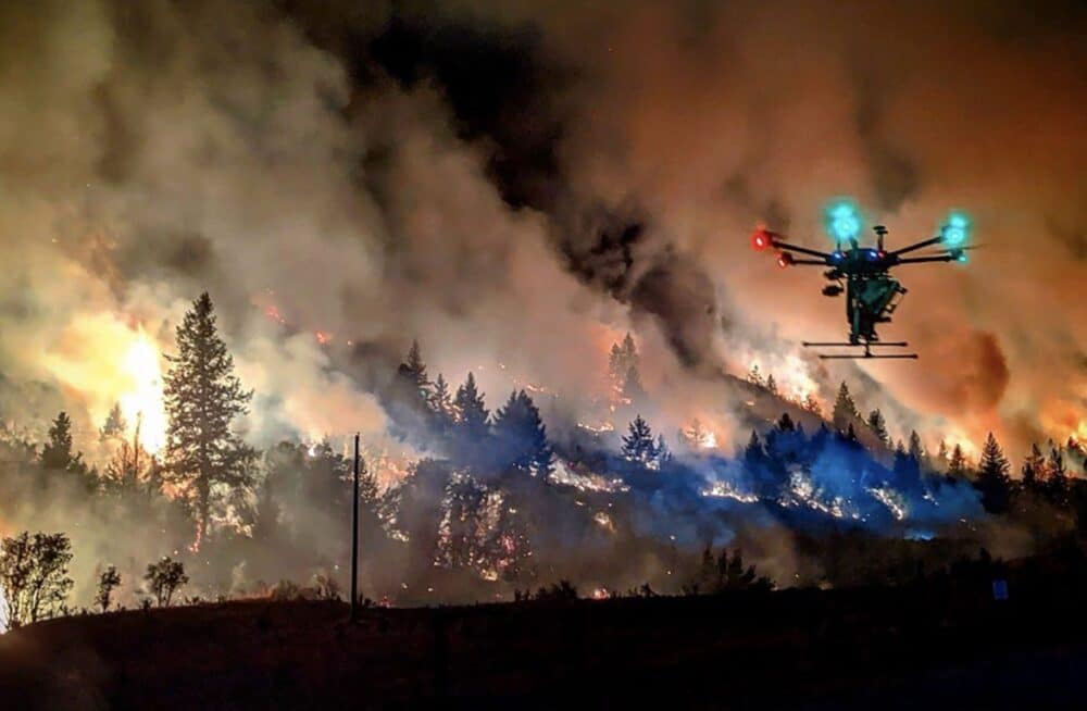 A multirotor drone flies past flames and smoke along a burning treeline.