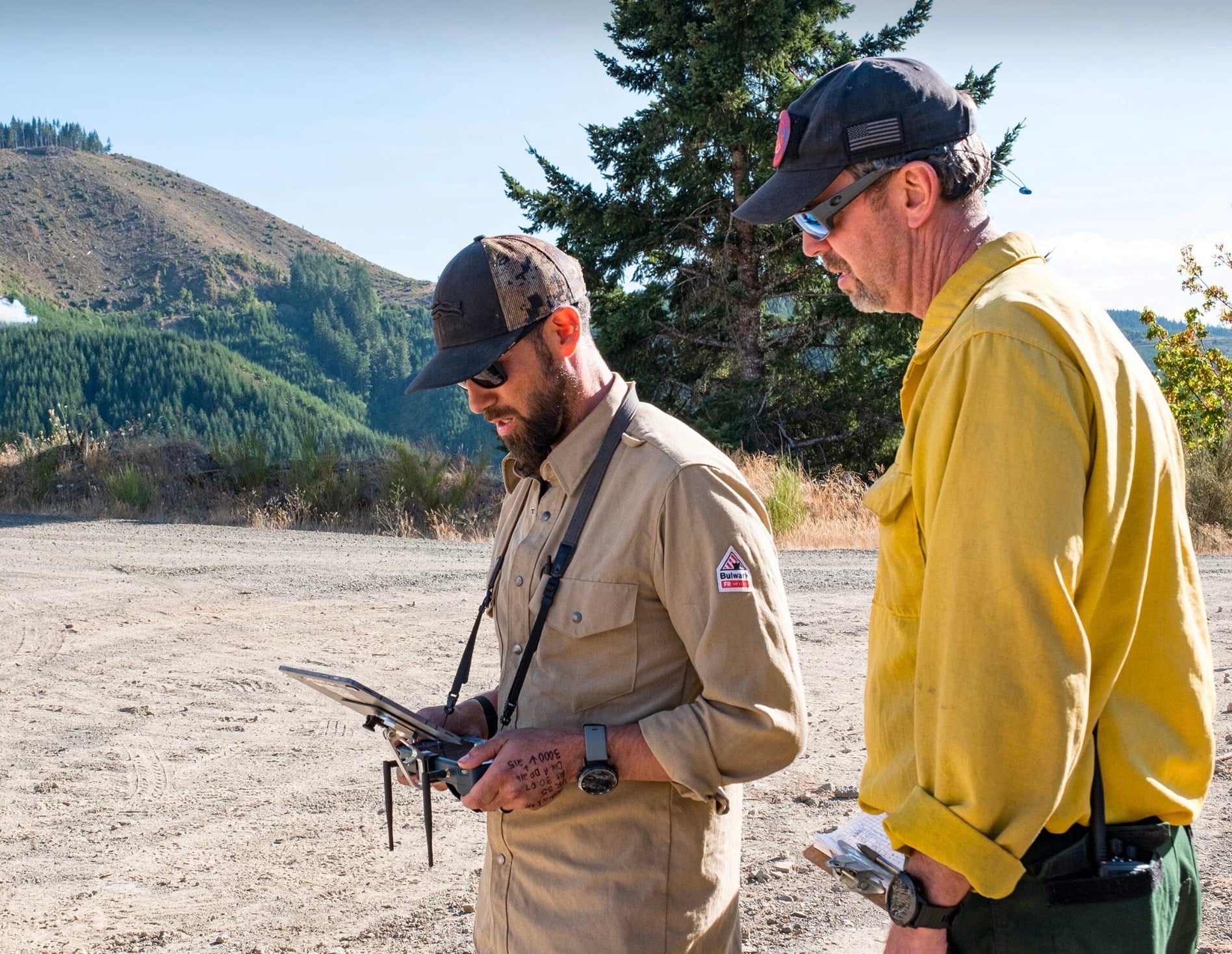 Two wildland firefighters study a drone controller on a gravel road below forested hills.