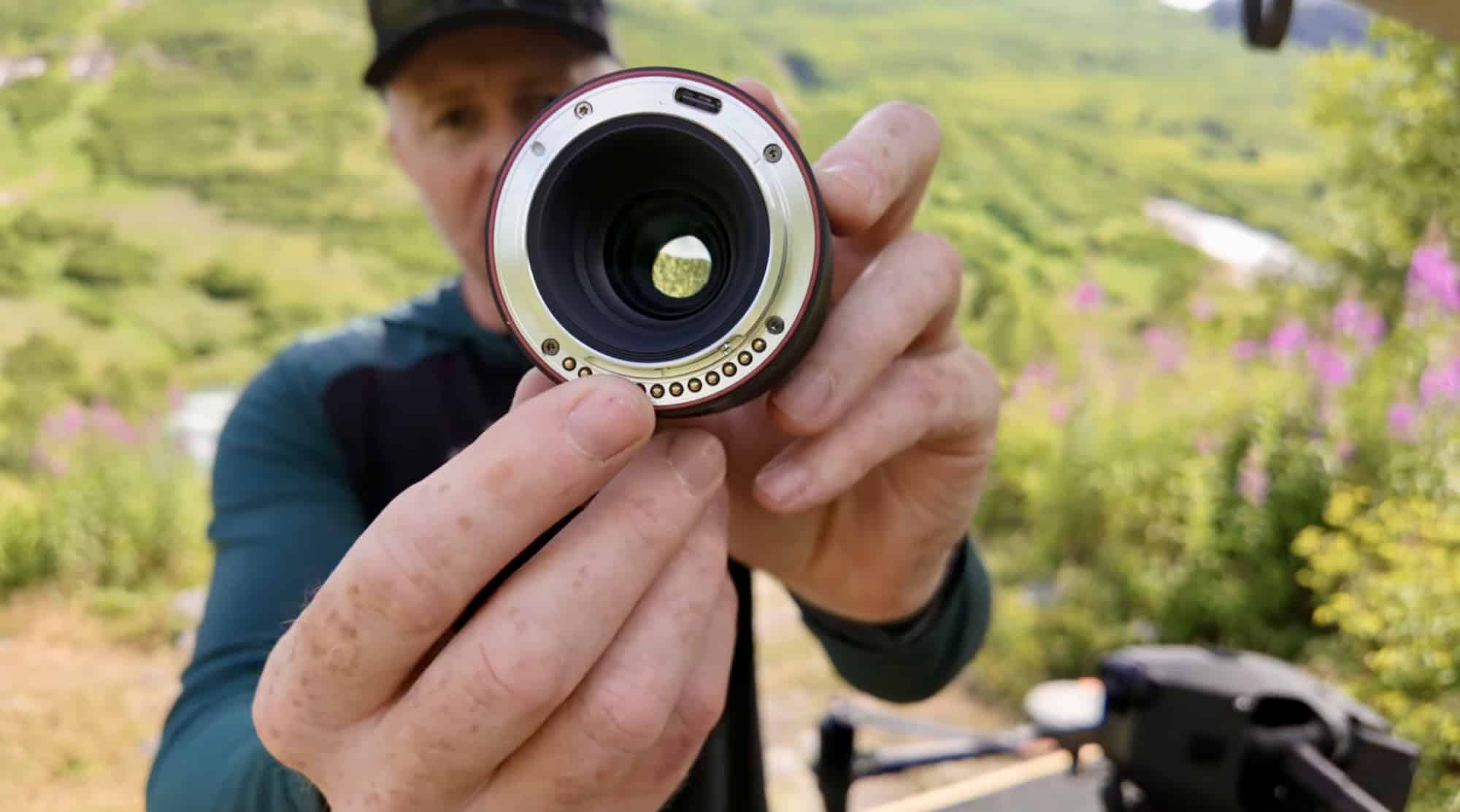 Presenter holding the 90mm DL lens toward camera in a green alpine valley.