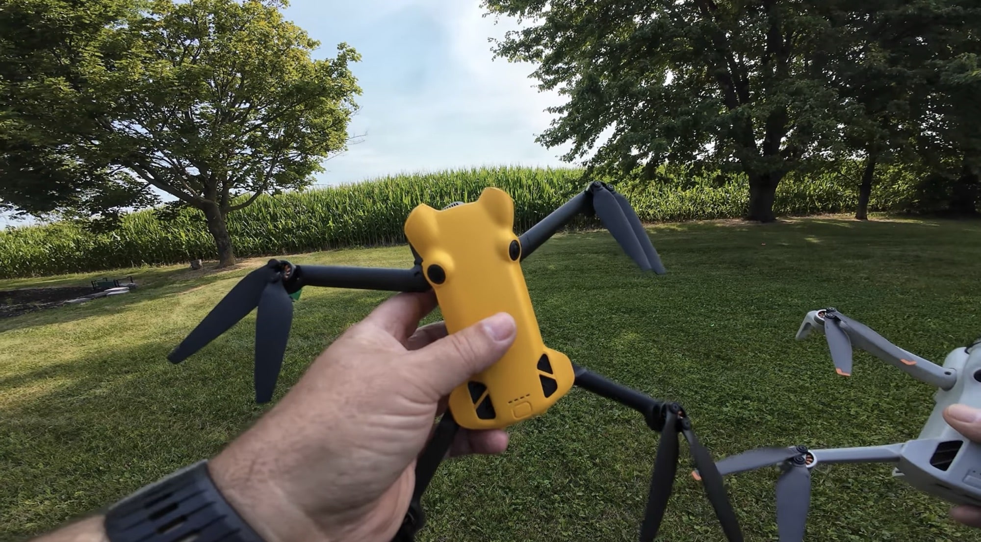 A hand holds the yellow Skyrover X1 above a lawn beside a cornfield.